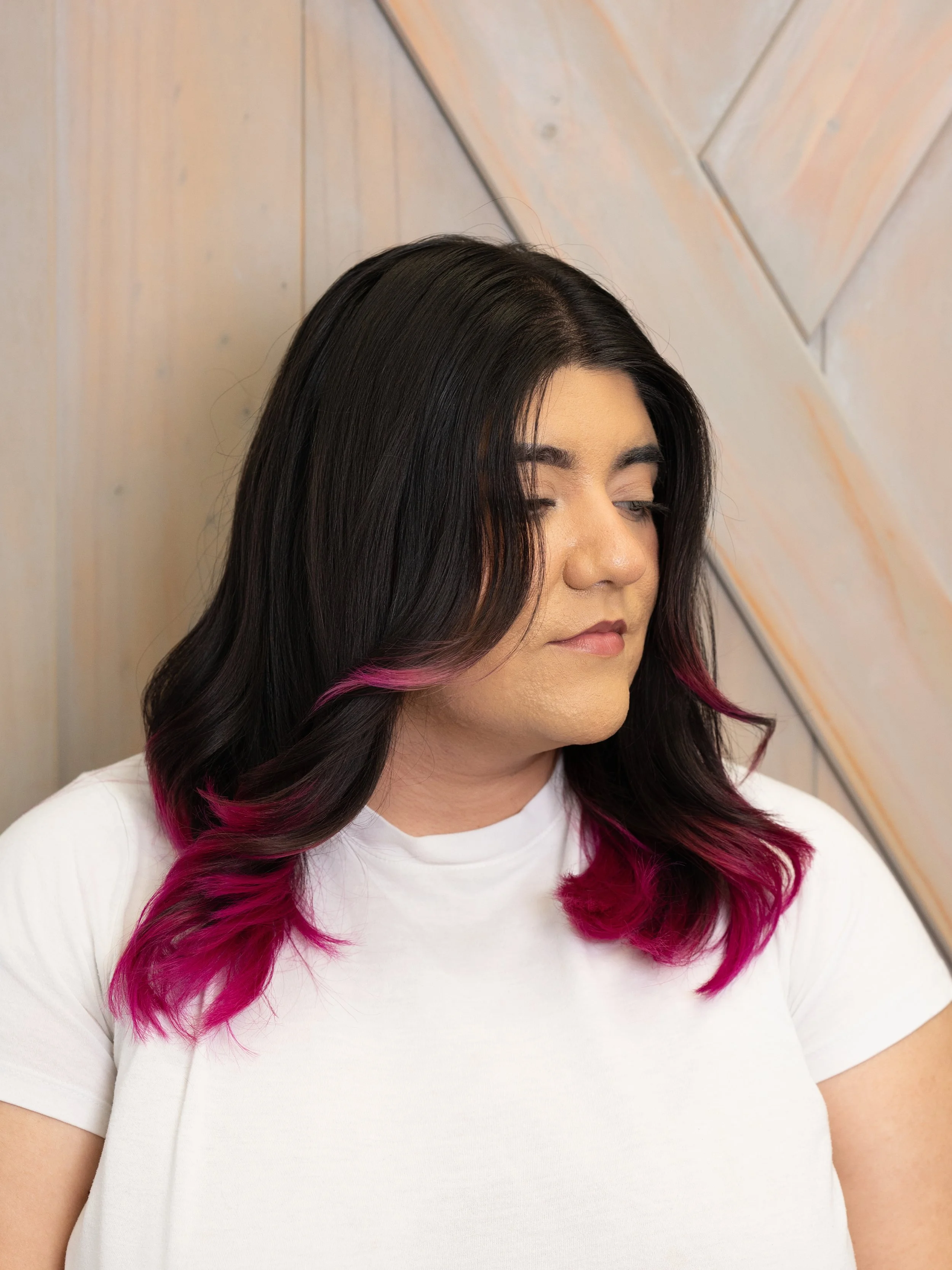 A young woman with long dark hair with pink ends, wearing a white shirt, standing in front of a wooden wall, looking downward with her eyes closed.