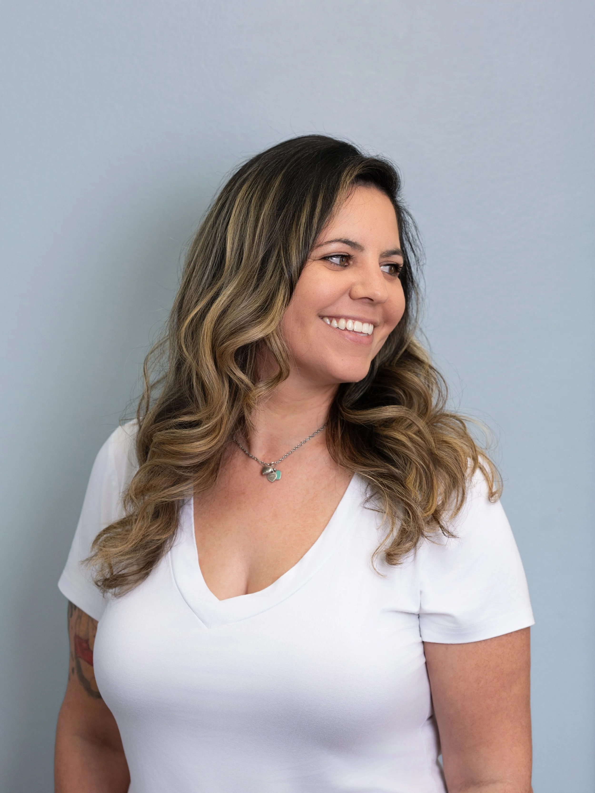 A woman with long wavy hair wearing a white V-neck shirt and a silver necklace, smiling, standing against a plain light grey wall.