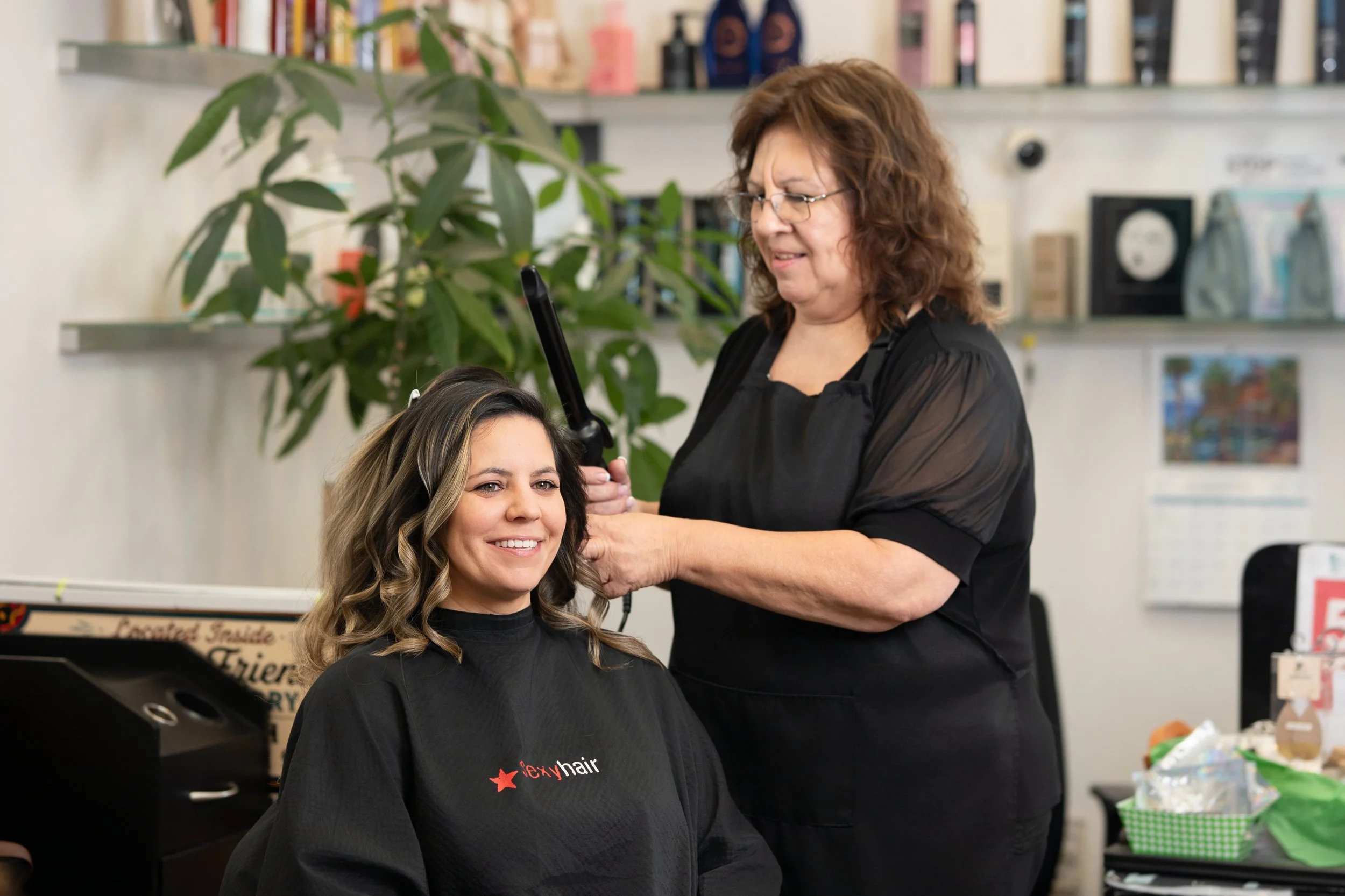 A hairstylist using a curling iron on a female client's hair in a salon. The woman is smiling and wearing a black cape with the logo 'alexy hair'.