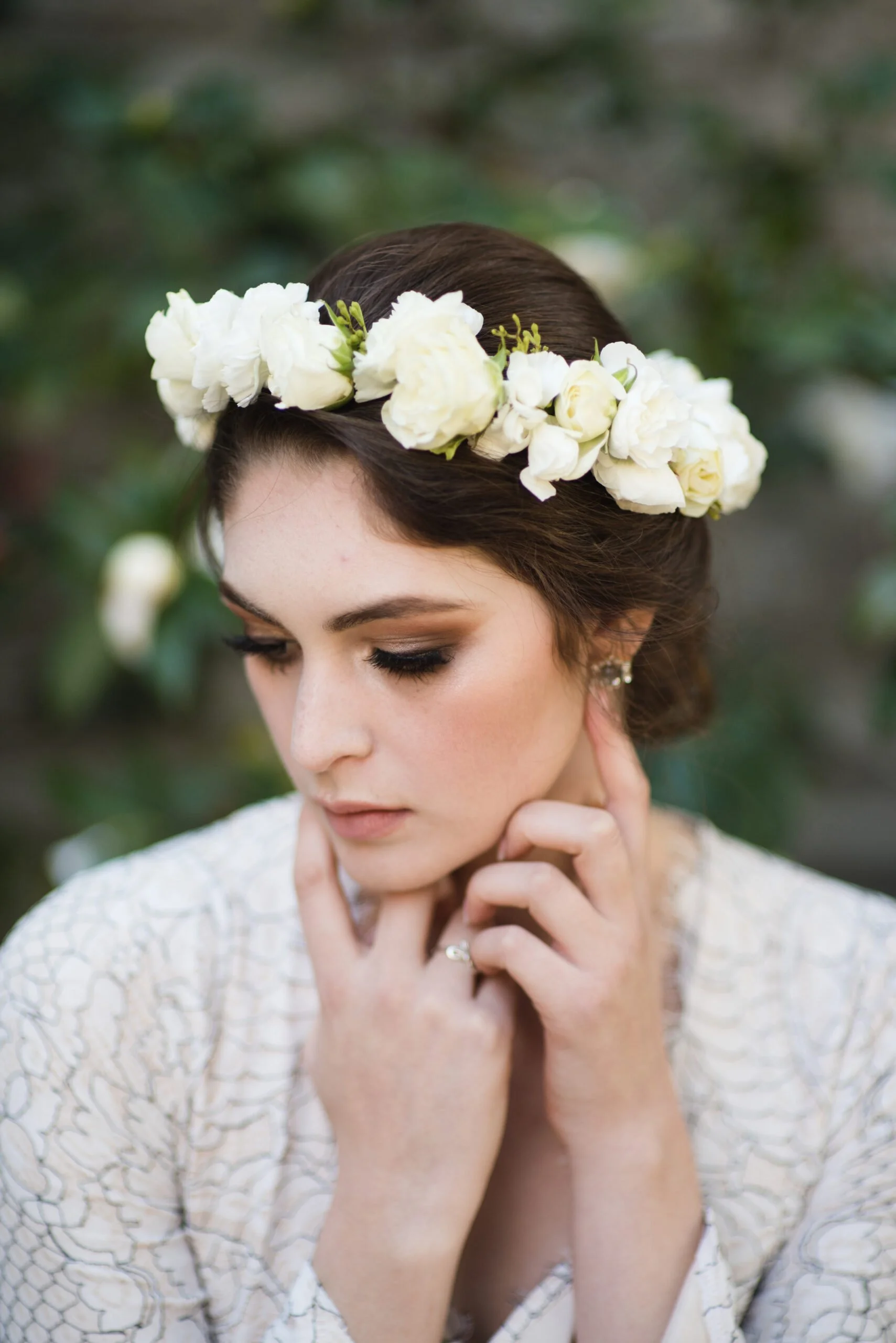 A woman with dark hair wearing a white floral crown and a white lace top, touching her face with fingers, with a blurred green natural background.