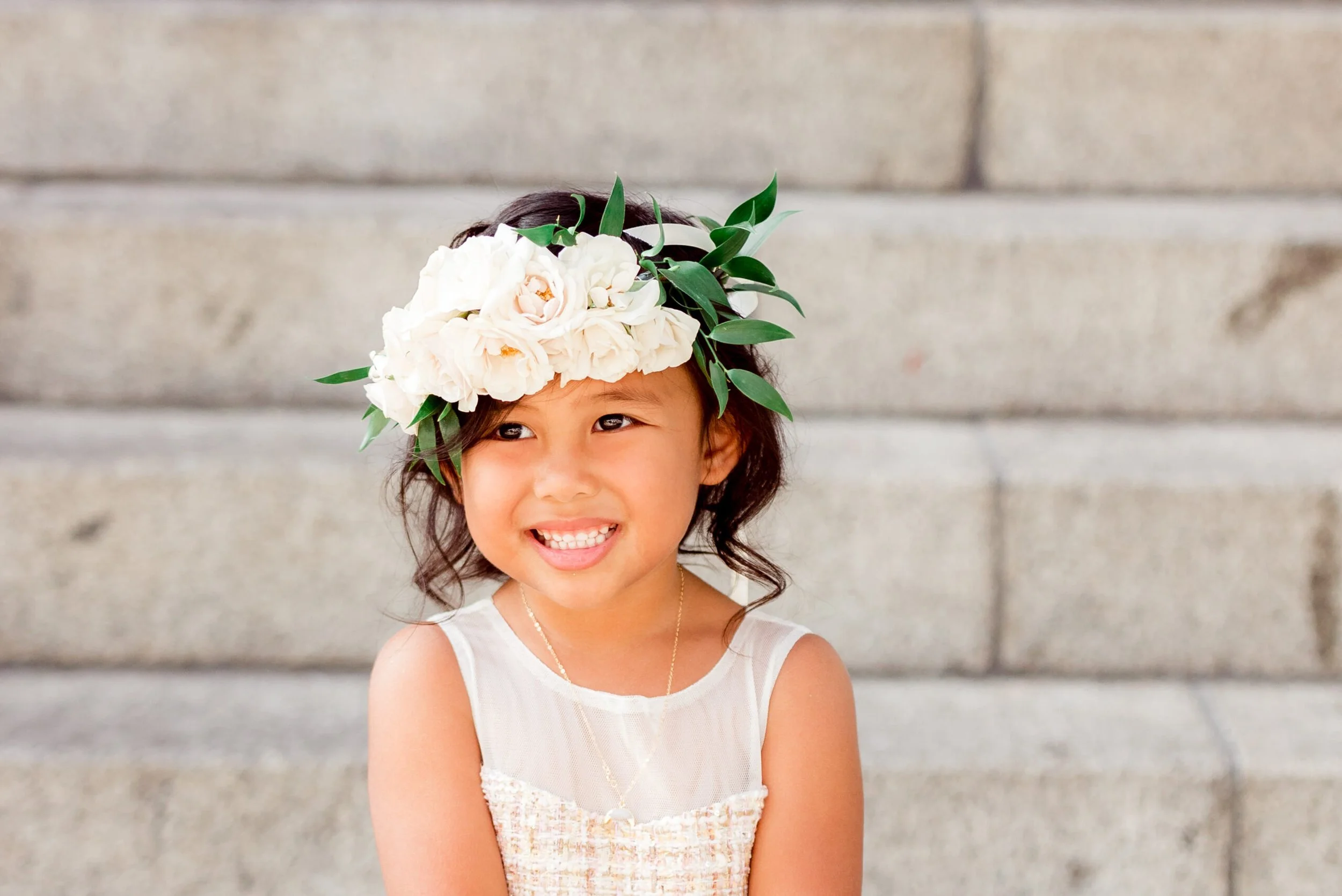 A young girl smiling, wearing a flower crown made of white flowers and green leaves, standing in front of a concrete wall.