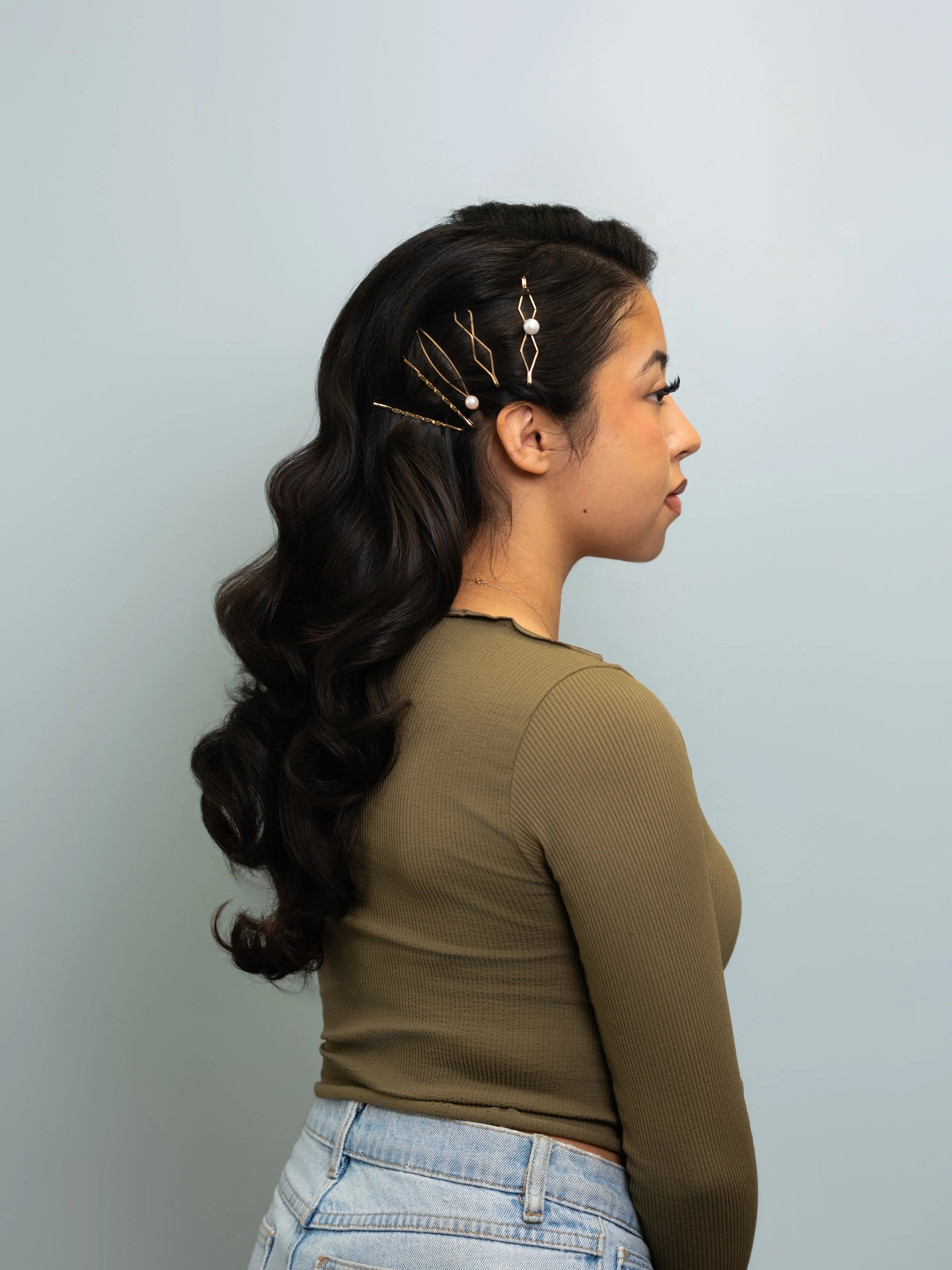 Side profile of a woman with wavy dark hair decorated with gold hairpins and pearl accents, wearing a khaki green top and light blue jeans, against a plain pale blue background.