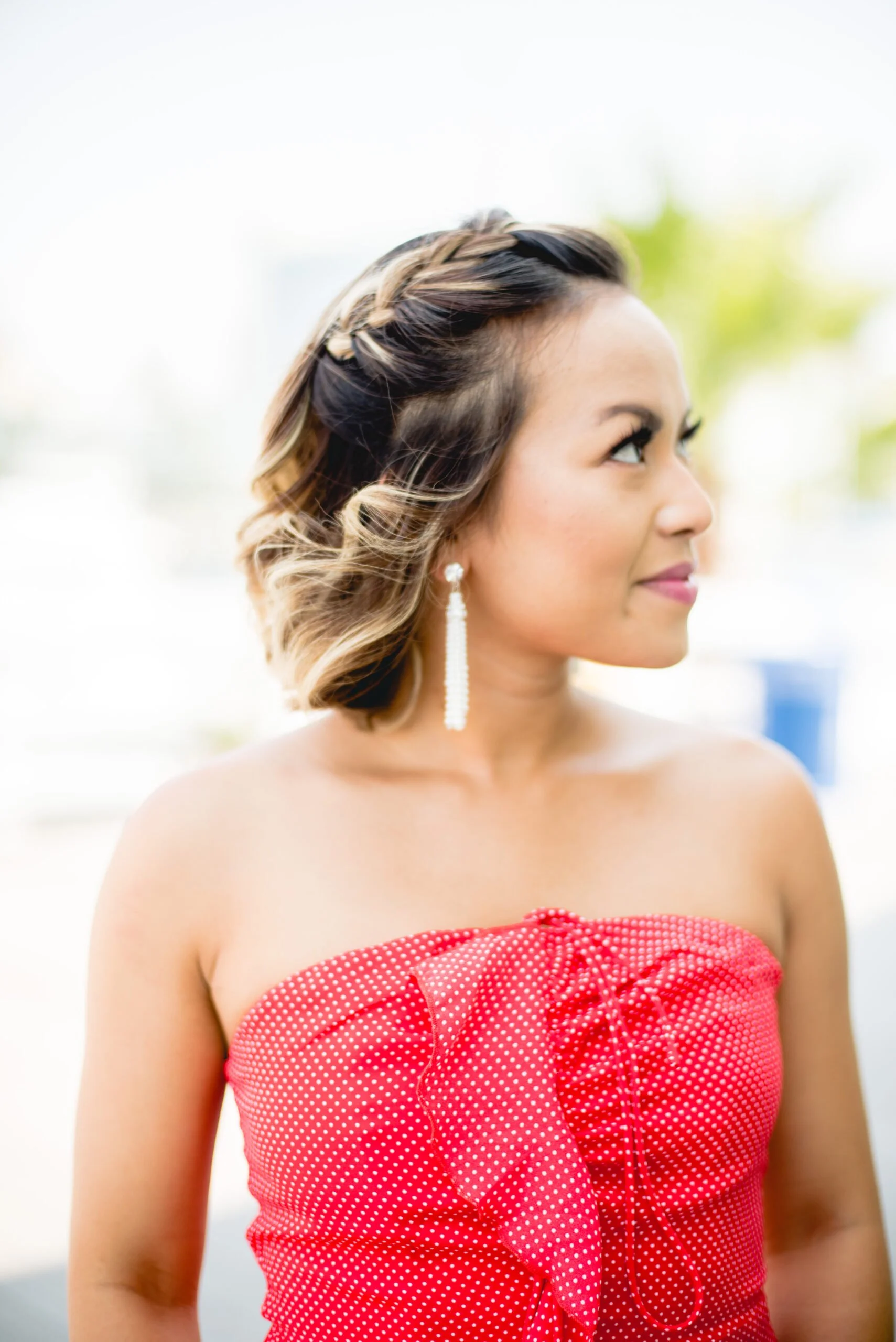 A woman with styled wavy hair wearing a strapless red dress with white polka dots, and dangling earrings, looking to her right outside.