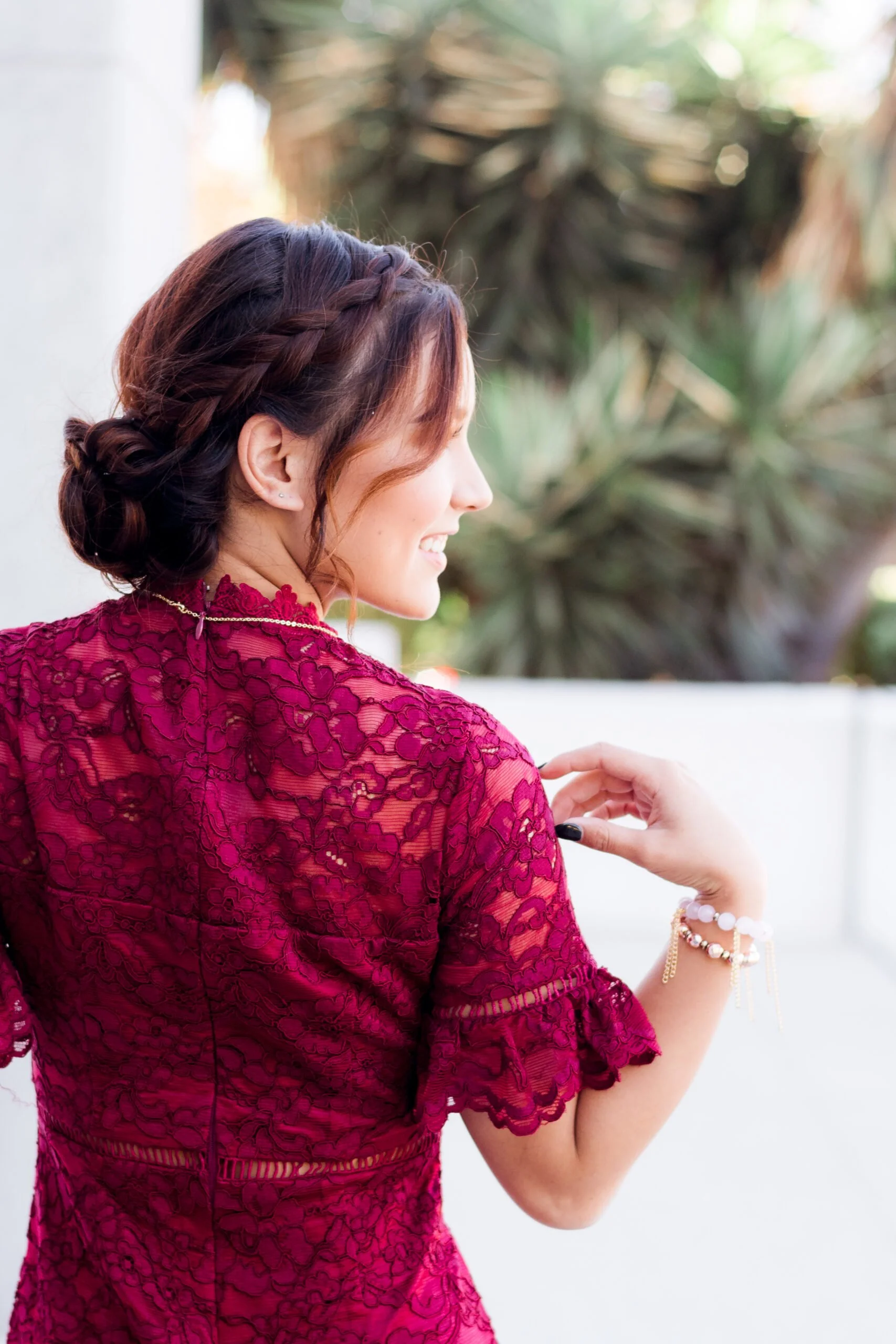 A woman with brown hair styled in braids and a bun, wearing a red lace dress with floral patterns, smiling and looking to the side, outdoors with blurred green plants in the background.