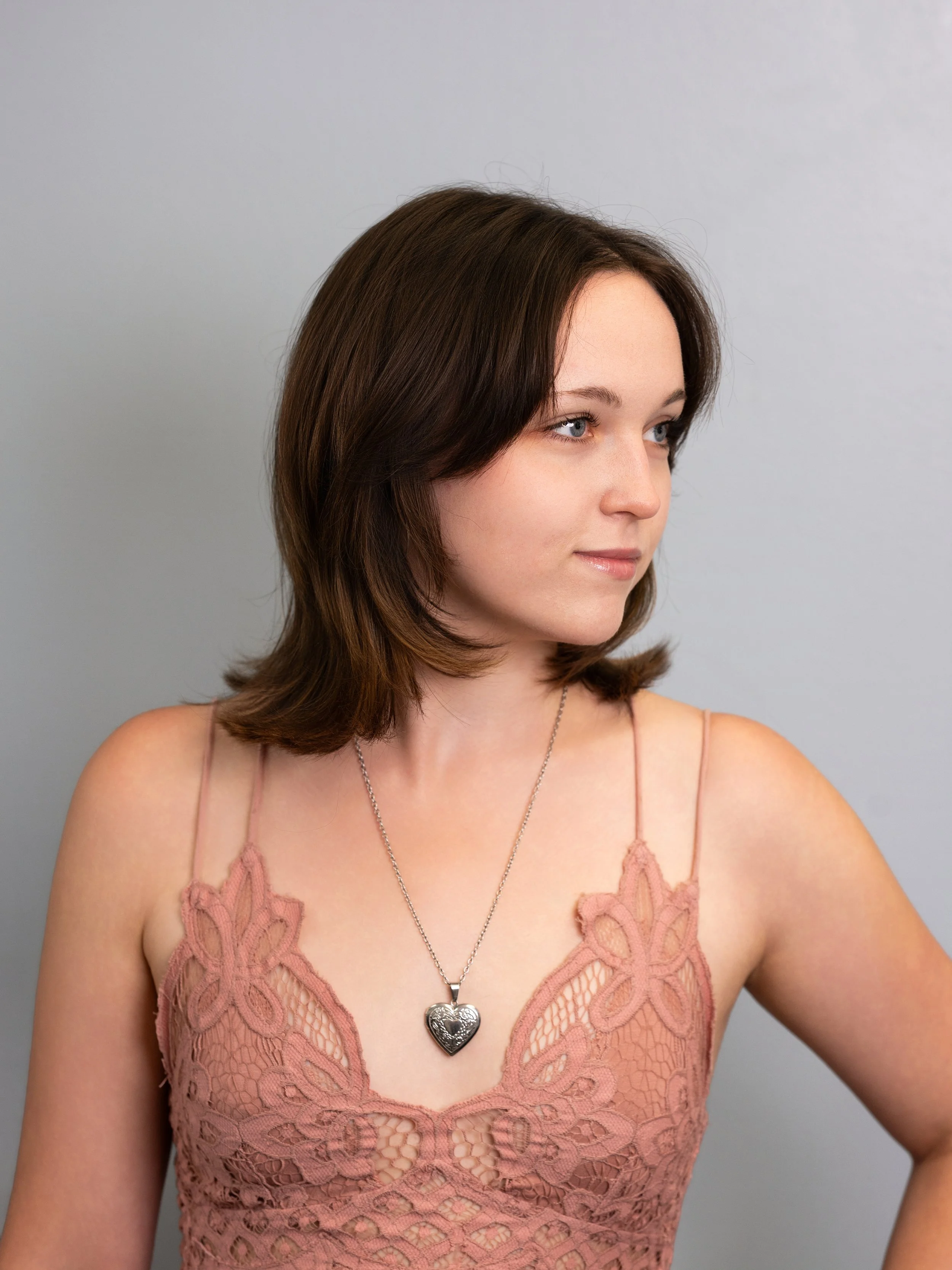 A woman with light skin and shoulder-length brown hair, wearing a pink lace top with spaghetti straps and a silver heart-shaped necklace, standing against a plain gray background.