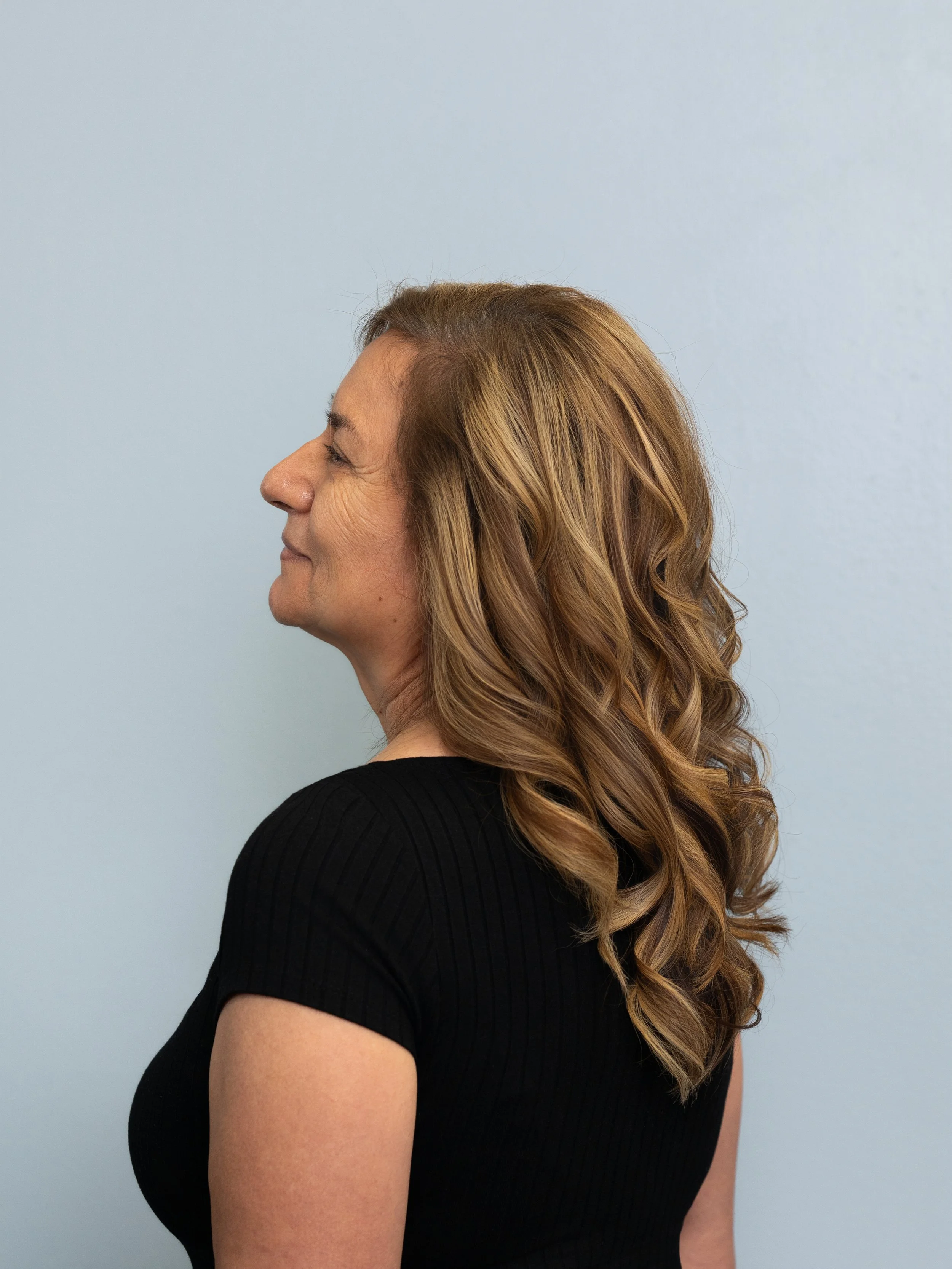 Side profile of a woman with wavy light brown hair wearing a black shirt against a light blue background.