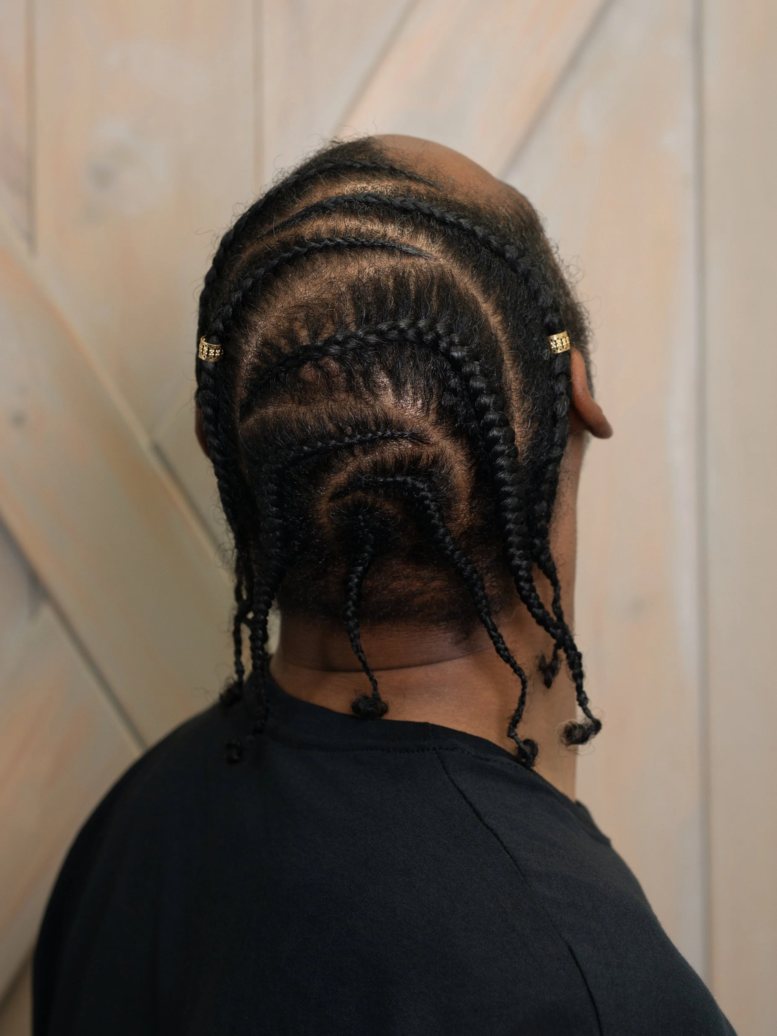 Back view of a person with braided hair, showing the scalp and hairline, wearing a black shirt with a wooden wall in the background.