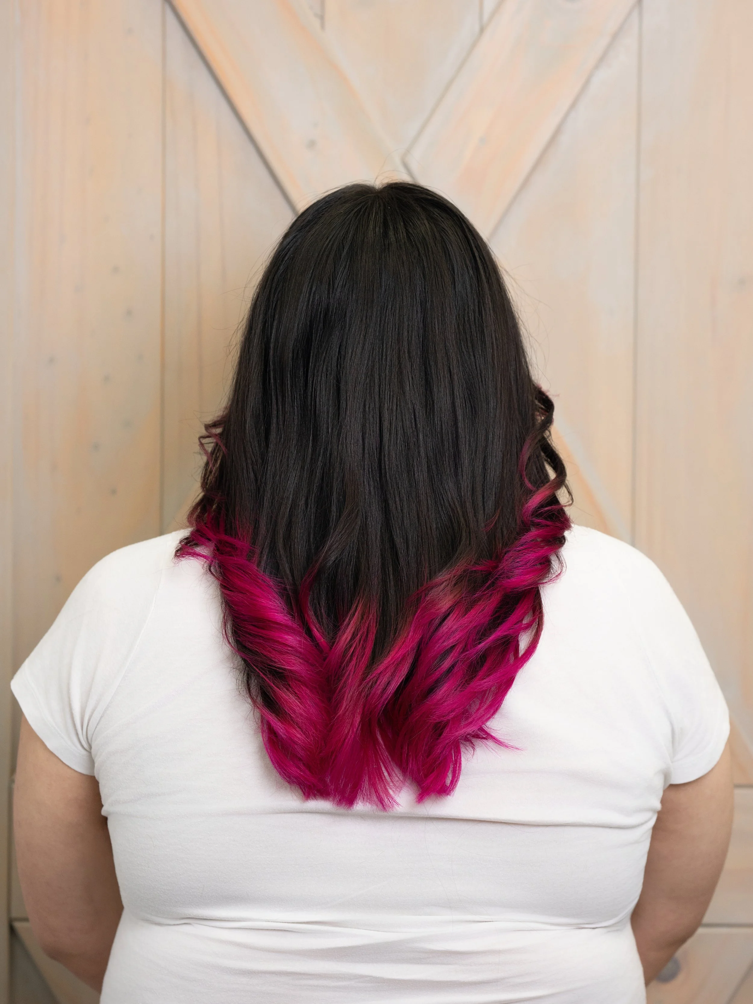 Back view of a woman with dark hair fading into bright pink at the tips, wearing a white t-shirt, standing against a wooden wall.