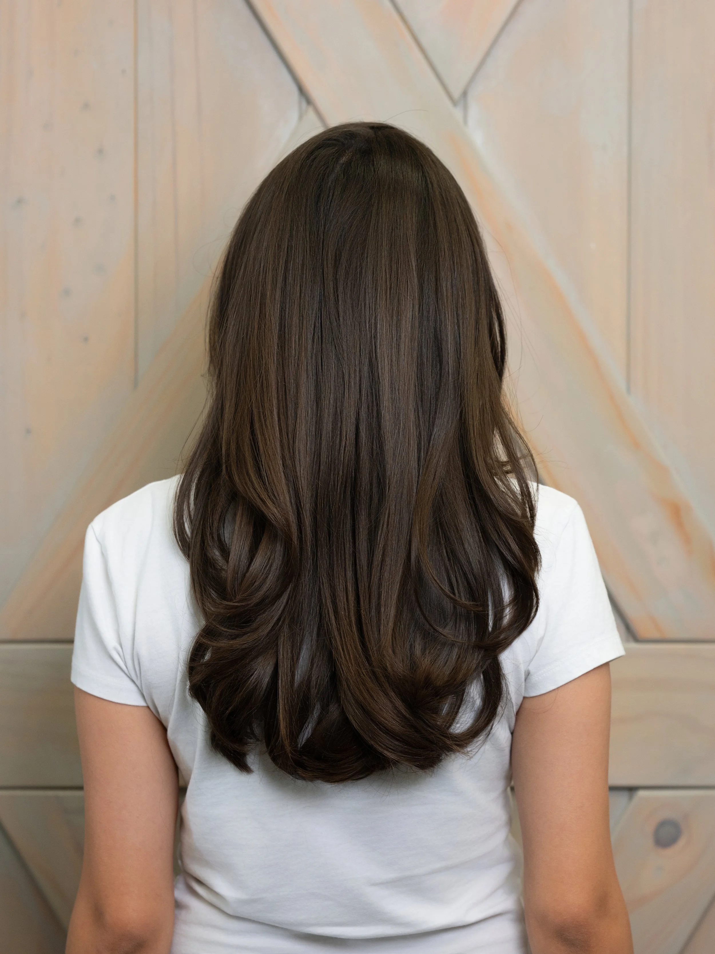 Back of a woman with long, brown, wavy hair wearing a white shirt, standing in front of a wooden wall with a geometric pattern.