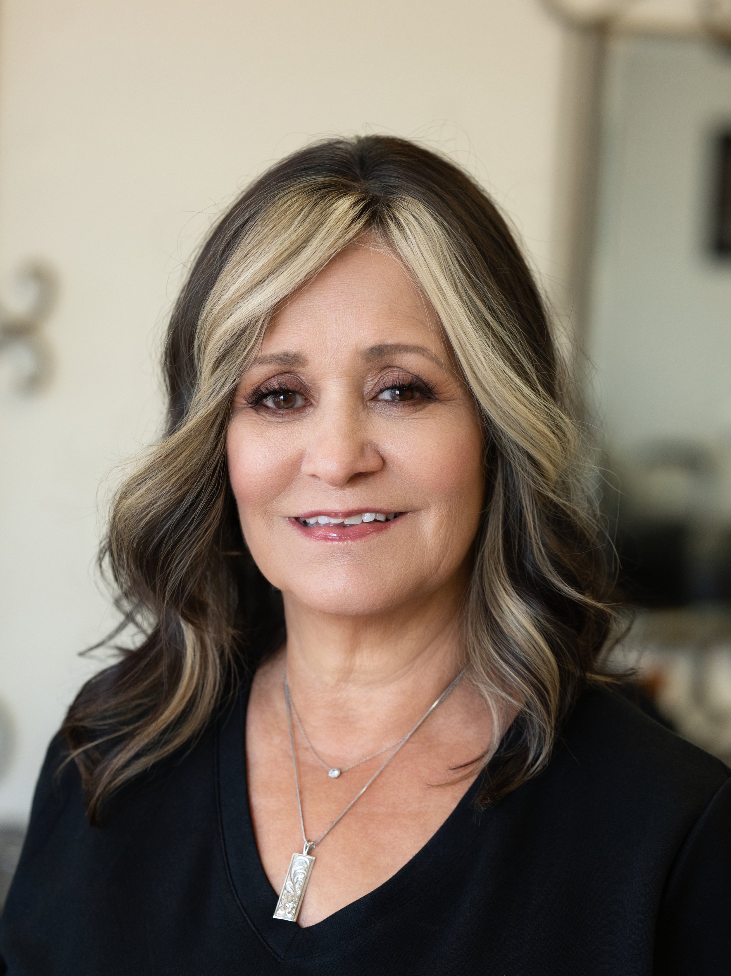 Close-up of a smiling woman with shoulder-length hair, wearing a black top, a silver necklace with a pendant, and a smaller chain with a round charm. The background is blurred.