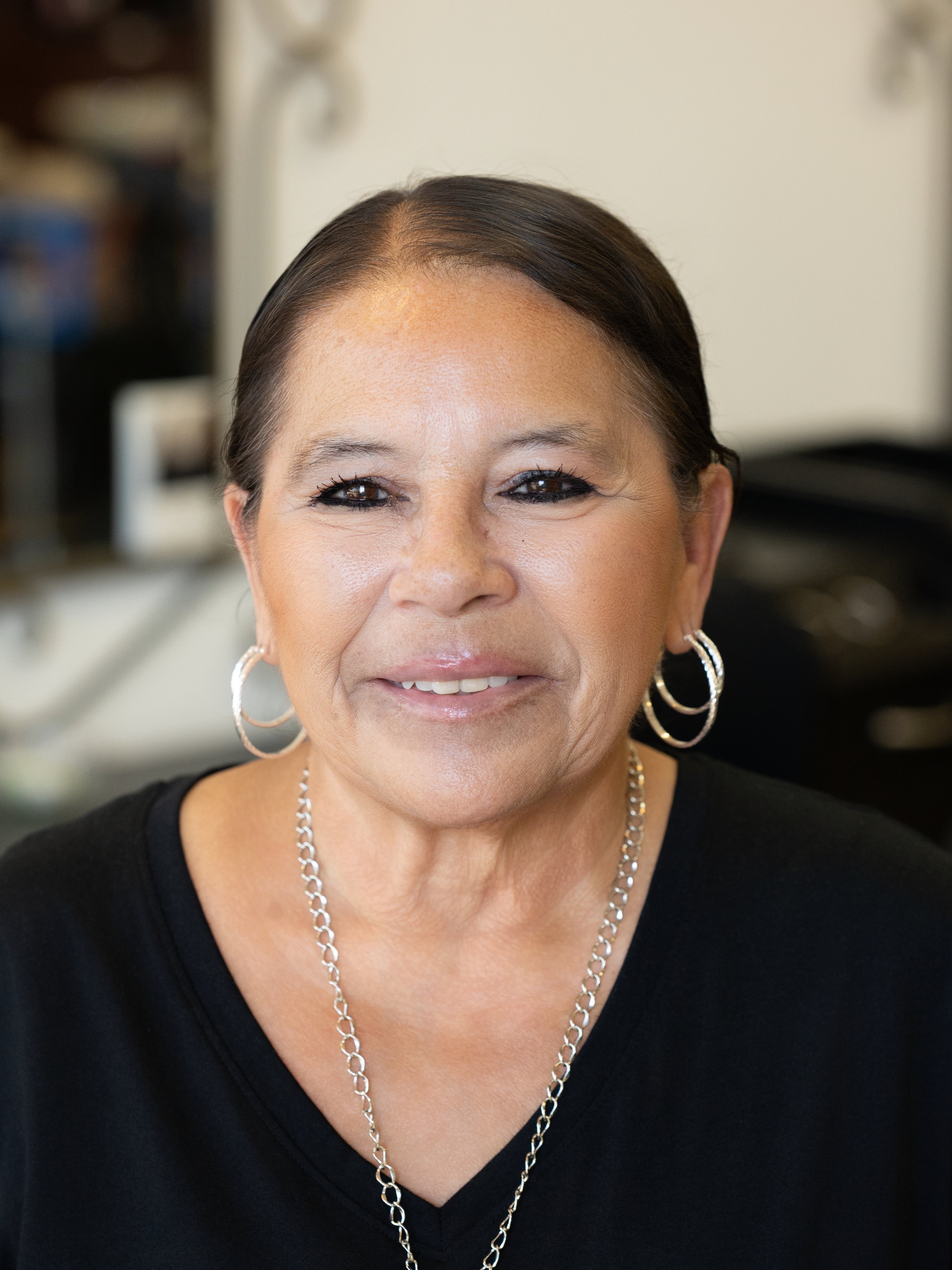 Close-up of a smiling woman with short dark hair, wearing hoop earrings and a silver chain necklace, in front of a blurred background.
