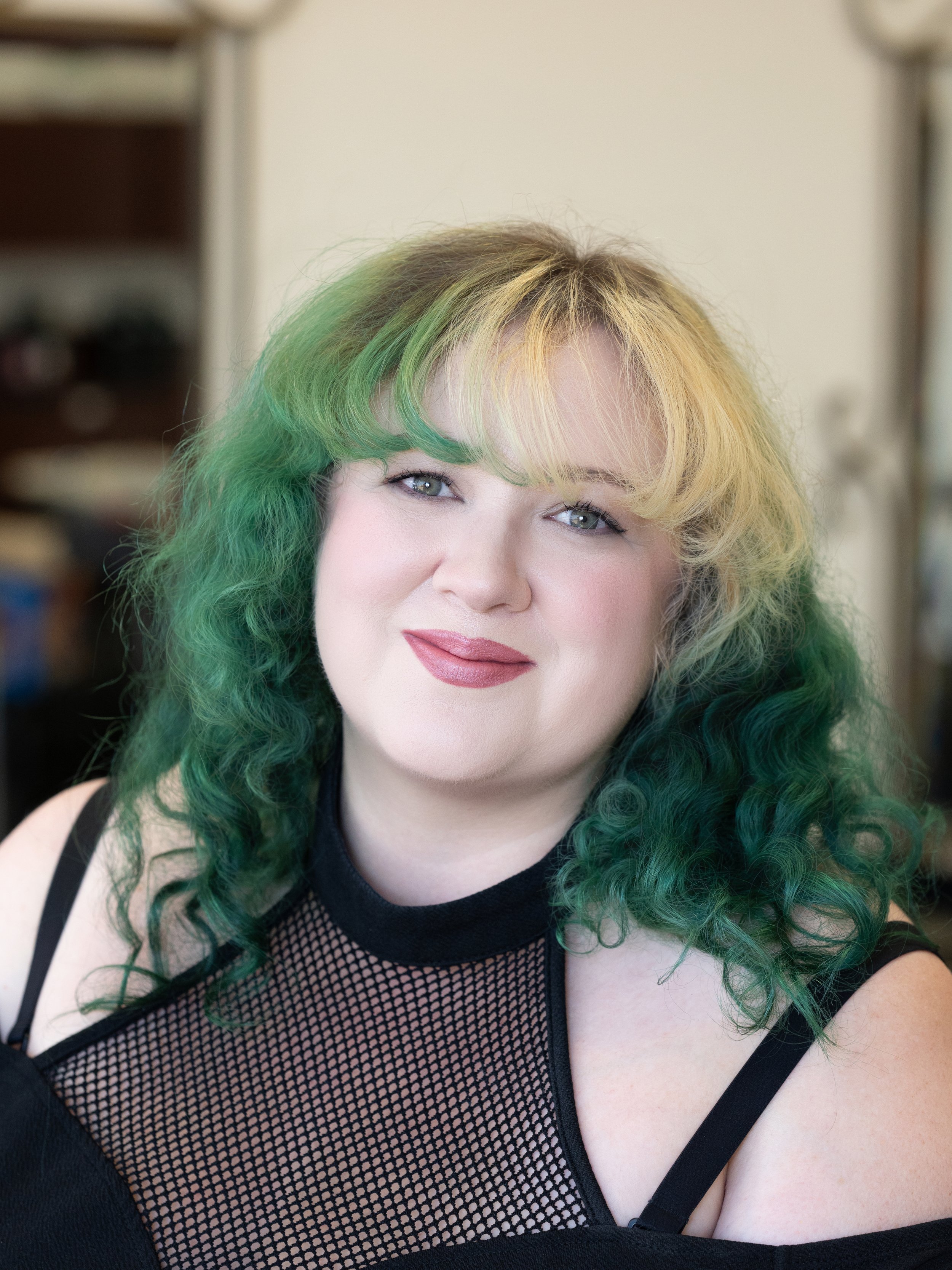 A woman with multicolored hair, primarily green and blonde, smiling at the camera. She is wearing a black sleeveless top with fishnet detail on the chest.