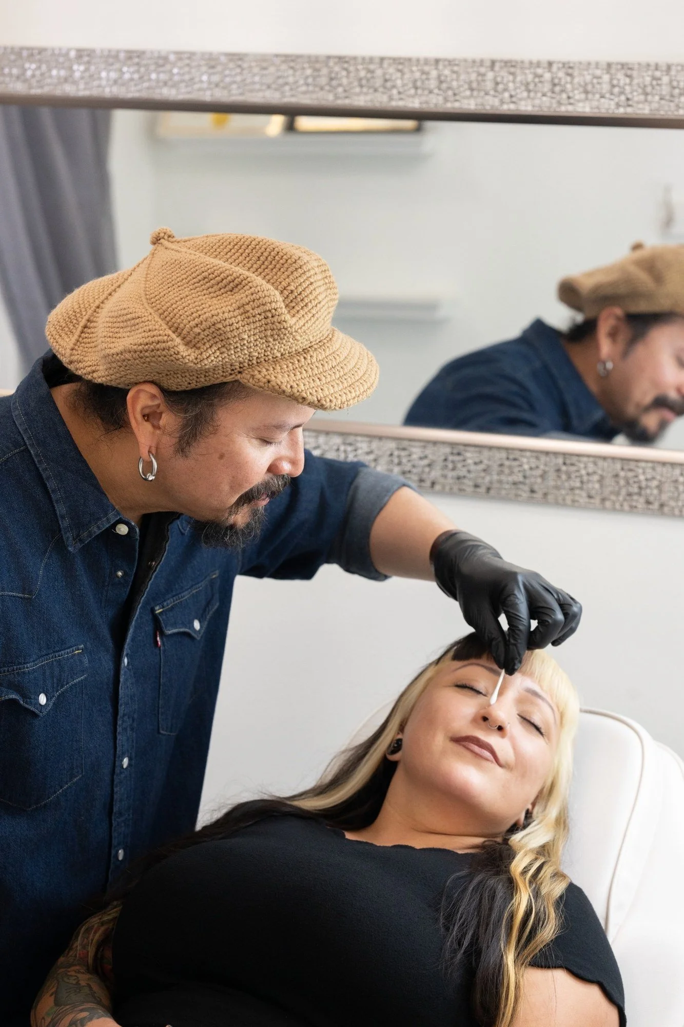 Tattooed woman with blonde hair receiving a cosmetic injection in her forehead from a practitioner wearing a beige knit beret and black gloves, with a mirror and decor in the background.