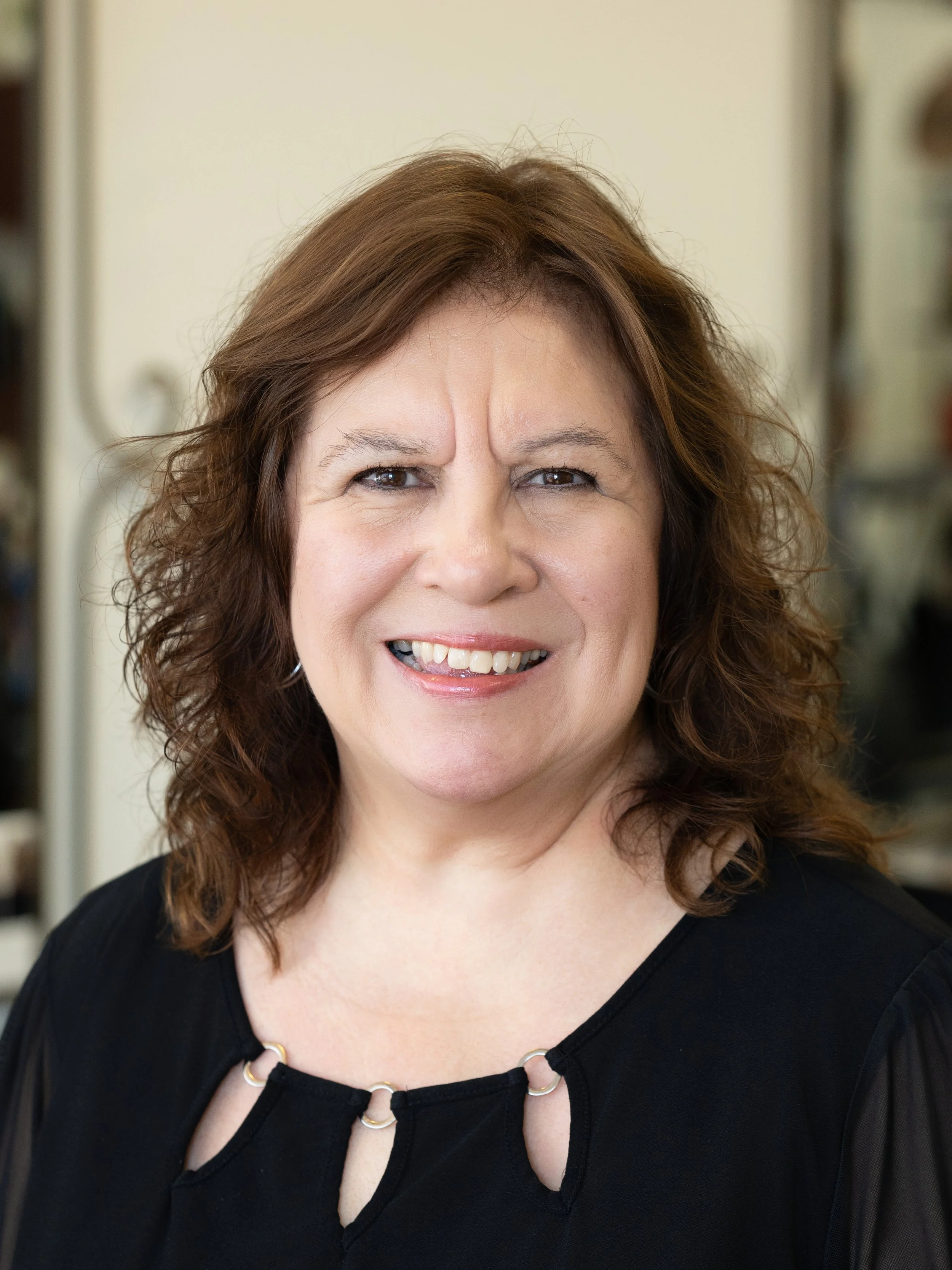 Mid-adult woman with brown, wavy hair smiling, wearing a black top and a necklace, in an indoor setting with shelves in the background.