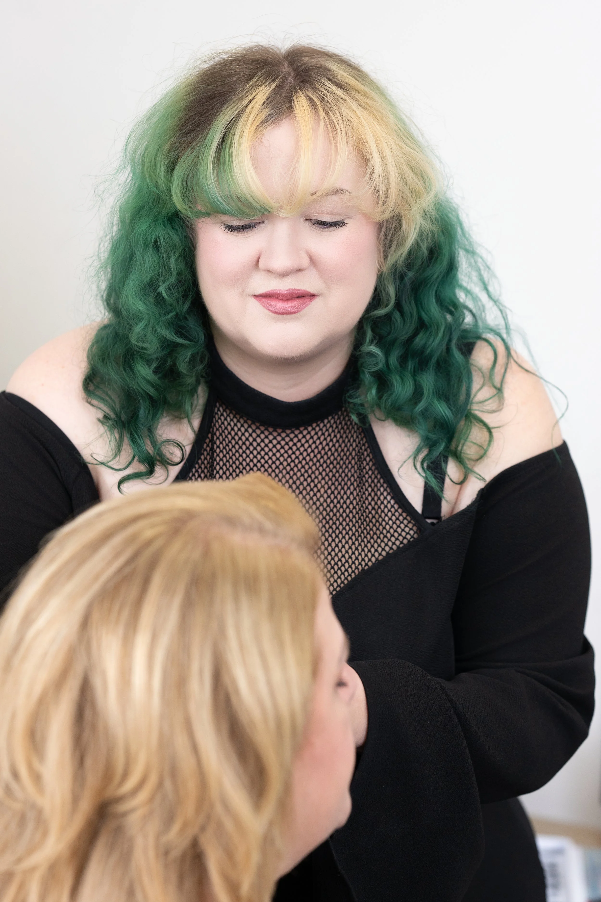 A woman with green and blonde curly hair, wearing a black mesh top and black off-shoulder clothing, is looking down and smiling at a person with long blonde hair.