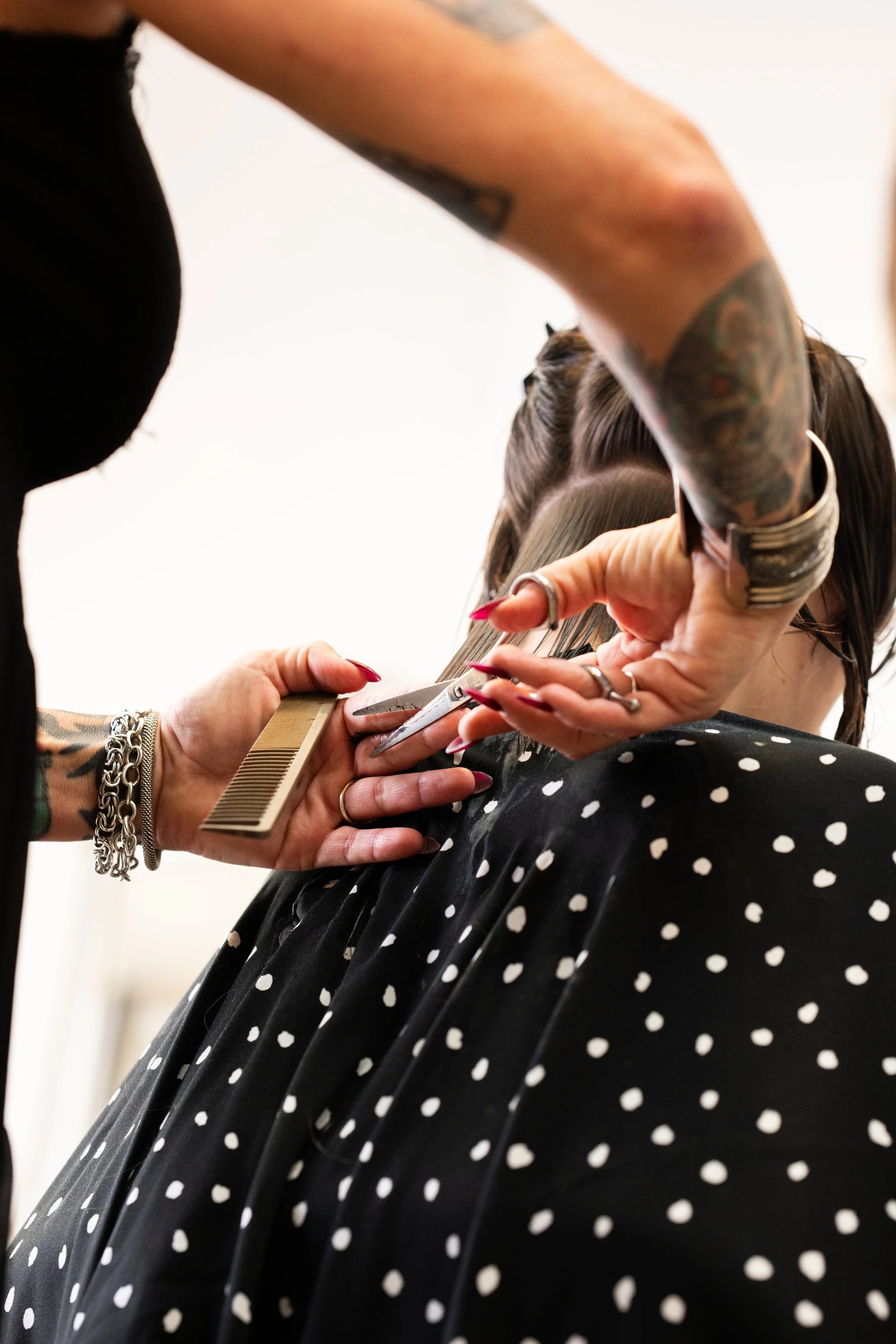 A hairstylist is trimming a seated client's hair with scissors and a comb, wearing jewelry and tattoos, in a salon with a black and white polka dot cape.