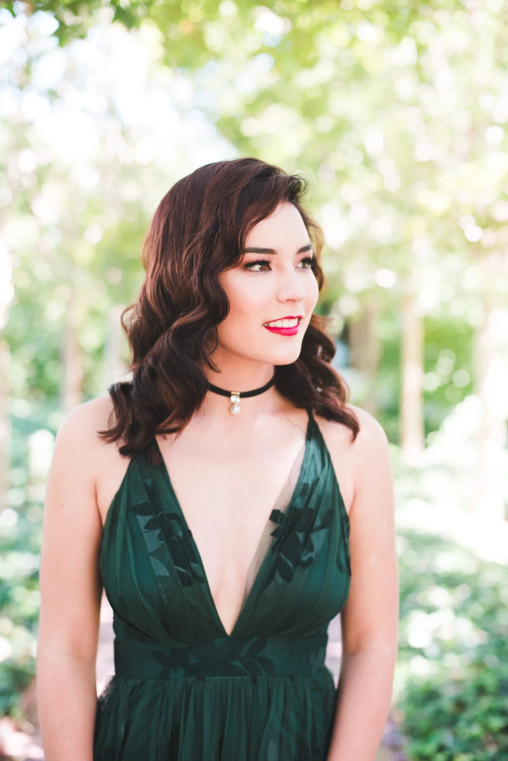 A woman with wavy dark hair and red lipstick wearing a black and green dress with a plunging neckline, outdoors with a background of green trees and sunlight.