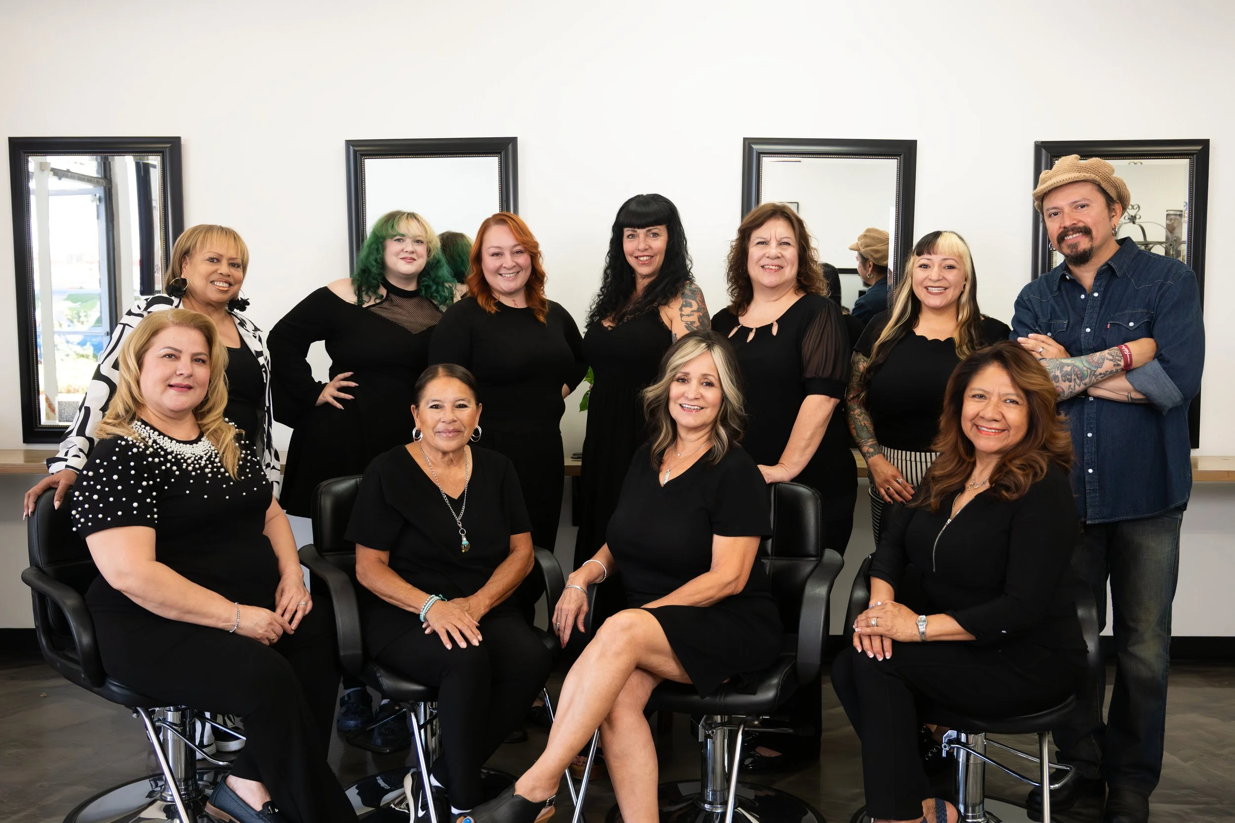 A group of ten diverse adults, mostly women, smiling and posing in a room with mirrors on the wall, some sitting and some standing, dressed in black and casual clothing.