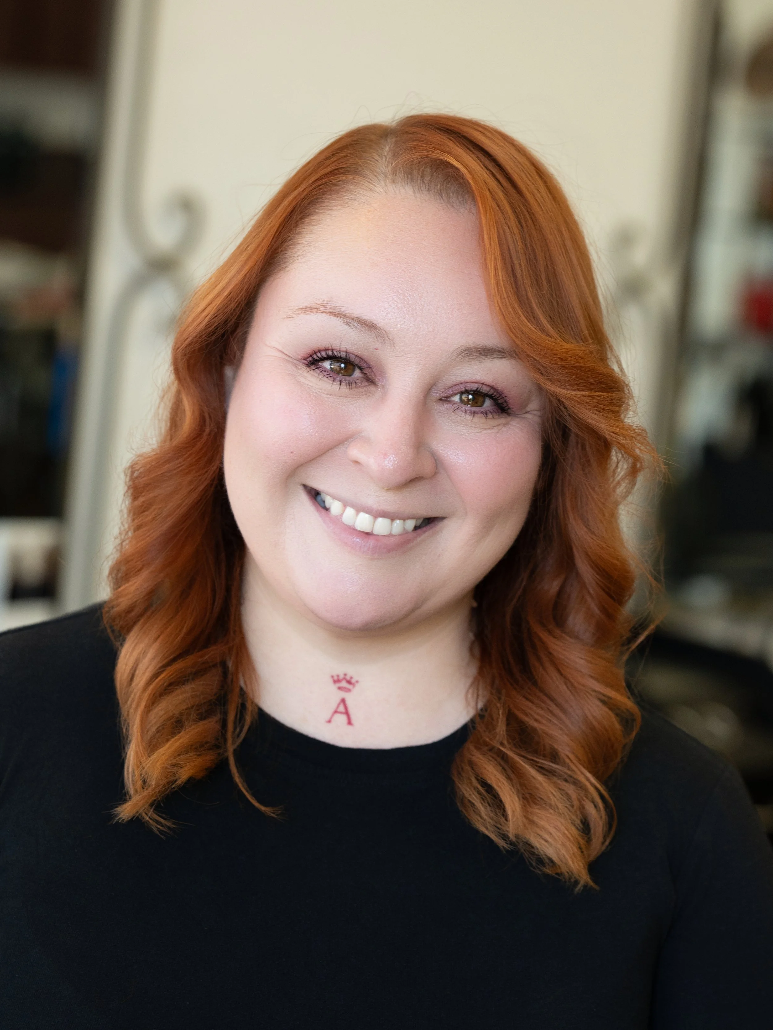 Close-up of a smiling woman with red wavy hair, wearing a black top, in an indoor setting.
