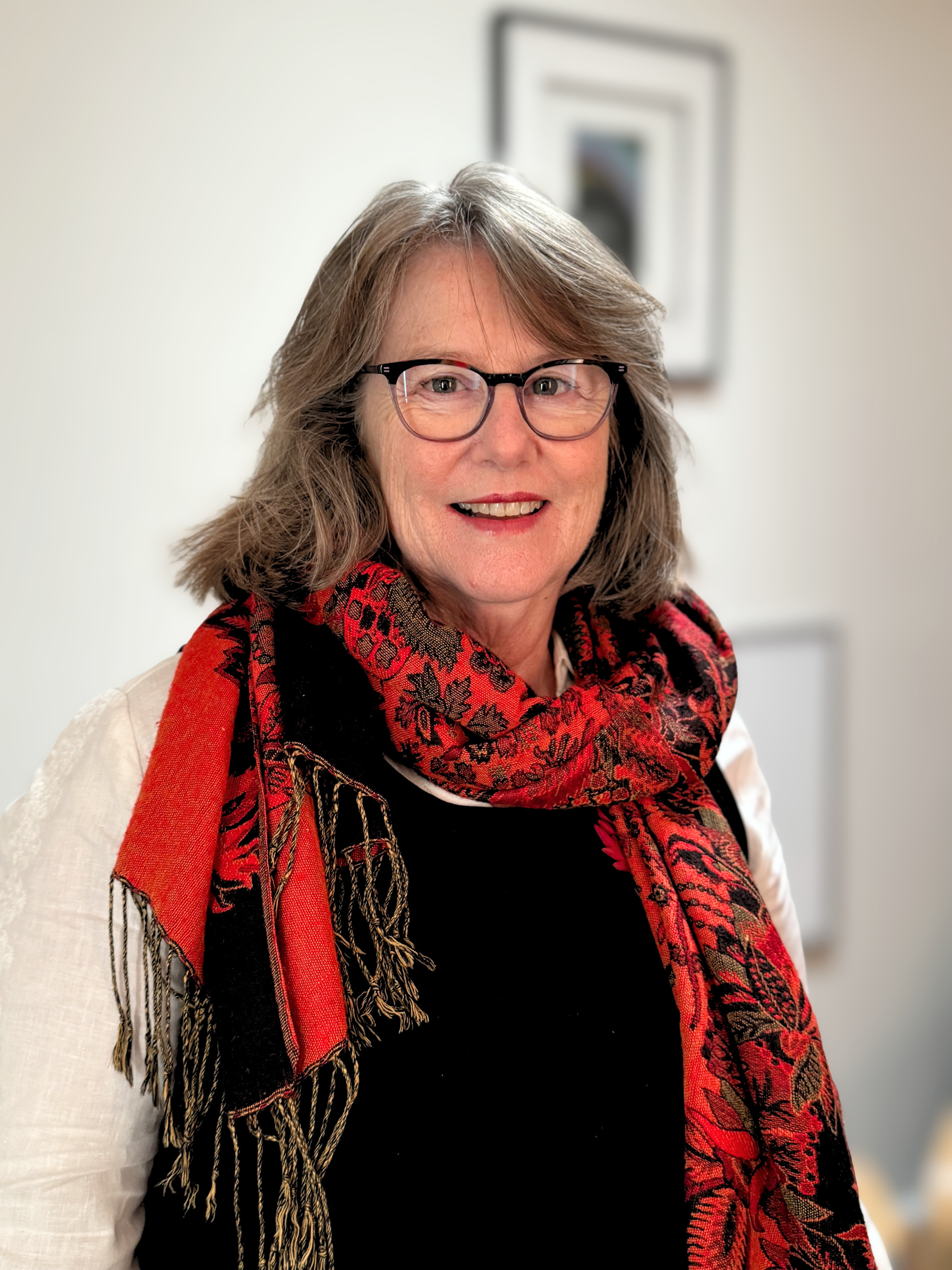 A middle-aged woman with shoulder-length brown hair wearing glasses, a black top, a red patterned scarf, and a white shirt with long sleeves, standing indoors in front of a blurred background with a framed picture on the wall.