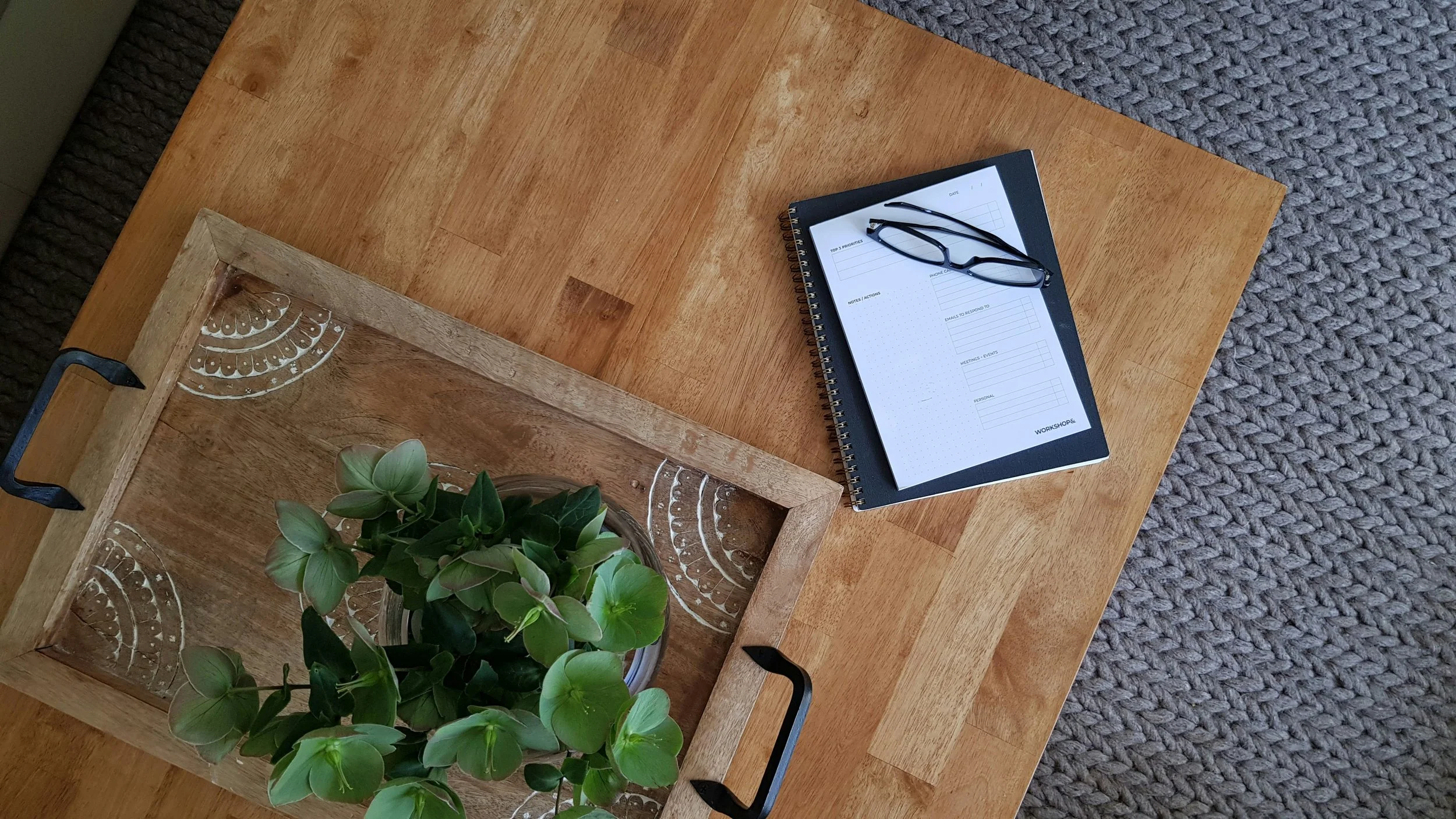 A wooden coffee table with a black notebook, a pair of black glasses, a potted green plant, a wooden tray with decorative white patterns, and a gray textured rug underneath.