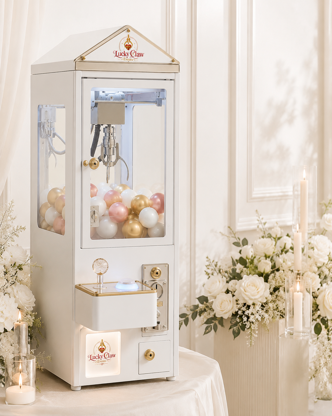 A white claw machine filled with pink, white, and gold balloons, situated on a table decorated with white flowers and candles in a softly lit, elegant room.