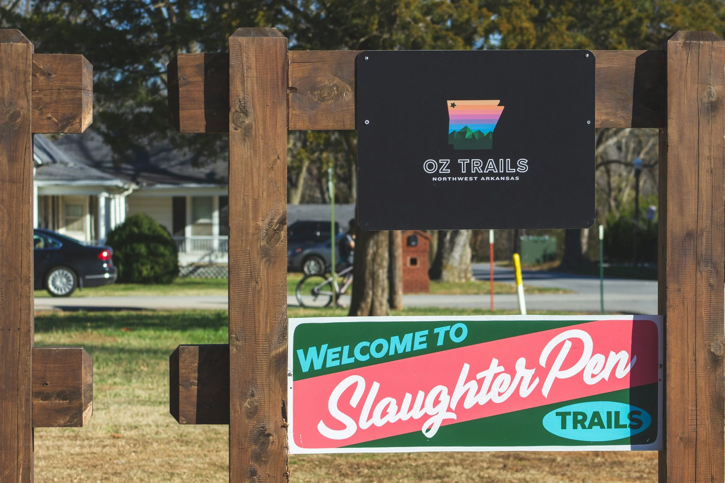 Sign for Oz Trails in Northwest Arkansas mounted on a wooden fence, with trees, houses, and cars visible in the background.