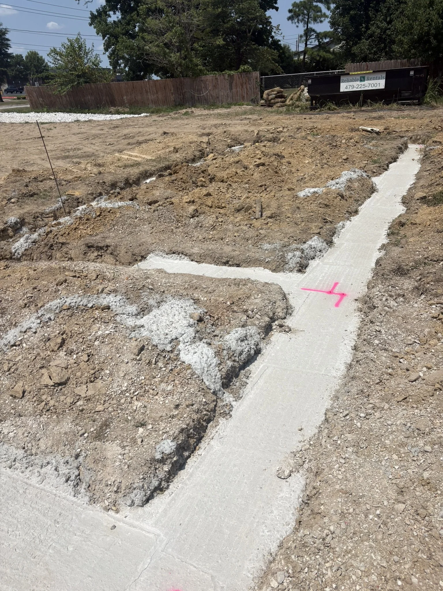 Construction site with dug-up soil, a newly poured concrete sidewalk, and markings in pink spray paint. In the background, there are trees, a wooden fence, and construction equipment with a sign for rental services.