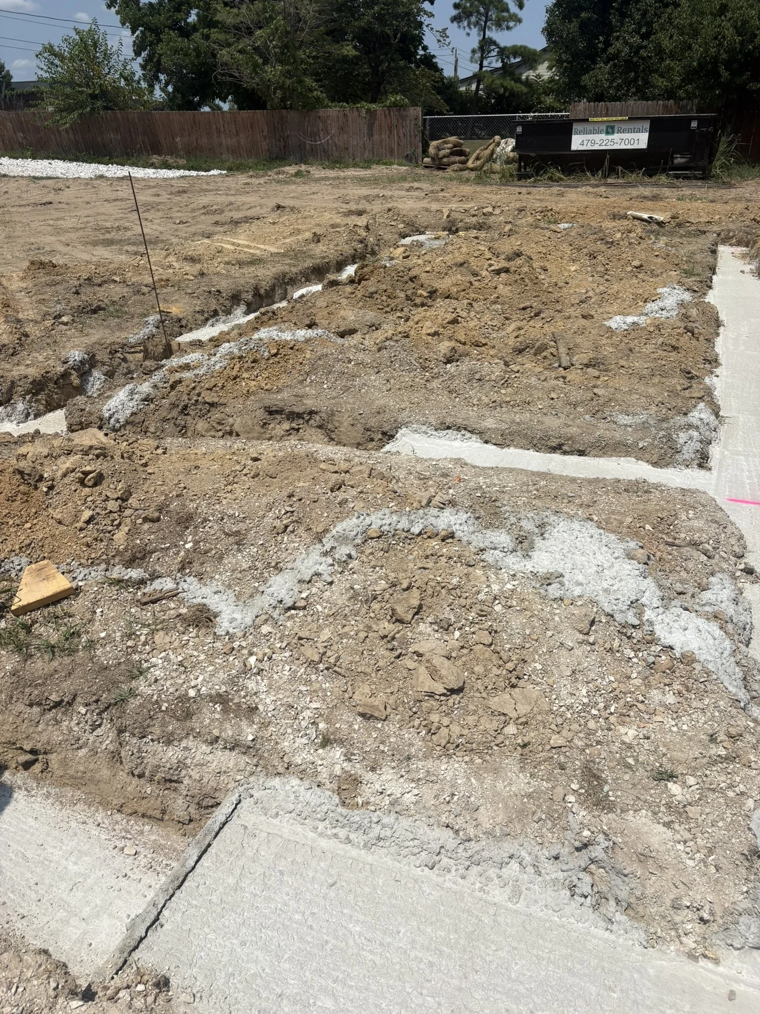 Construction site with excavated soil and a concrete sidewalk. In the background, there is a wooden fence, trees, a black dumpster, and some bags of gravel or sand.