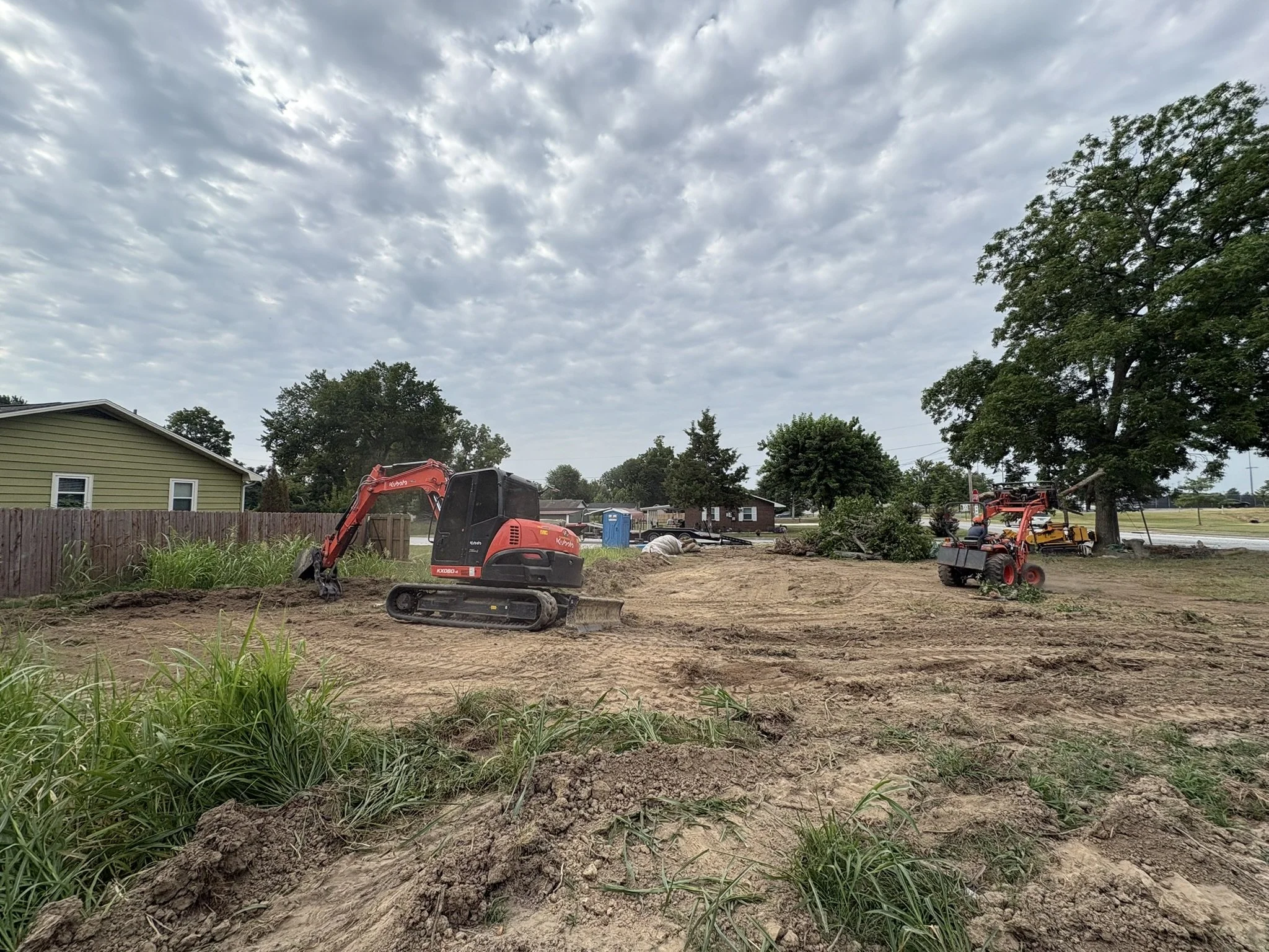 Construction site with dirt and grass, construction equipment such as a small excavator and a ride-on mower, residential houses in the background, cloudy sky overhead.
