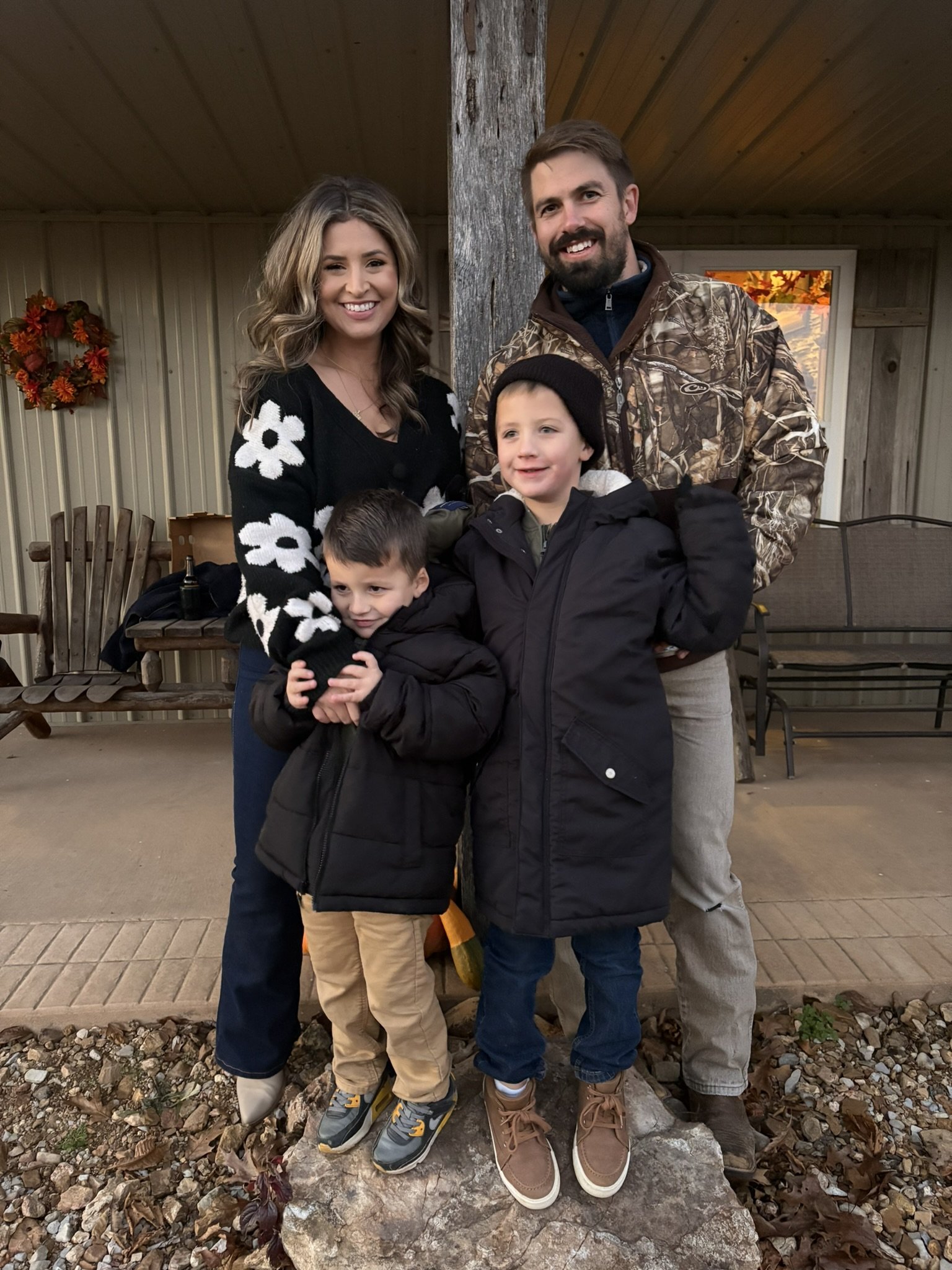 A family of four standing on a rock in autumn. The mother has wavy brown hair and is wearing a black sweater with white flowers. The father has short brown hair and a beard, wearing a camouflage jacket. The two children are boys wearing jackets; one has a black beanie and is smiling, the other has short hair and is looking down. The scene is outdoors with a wooden house and autumn leaves in the background.