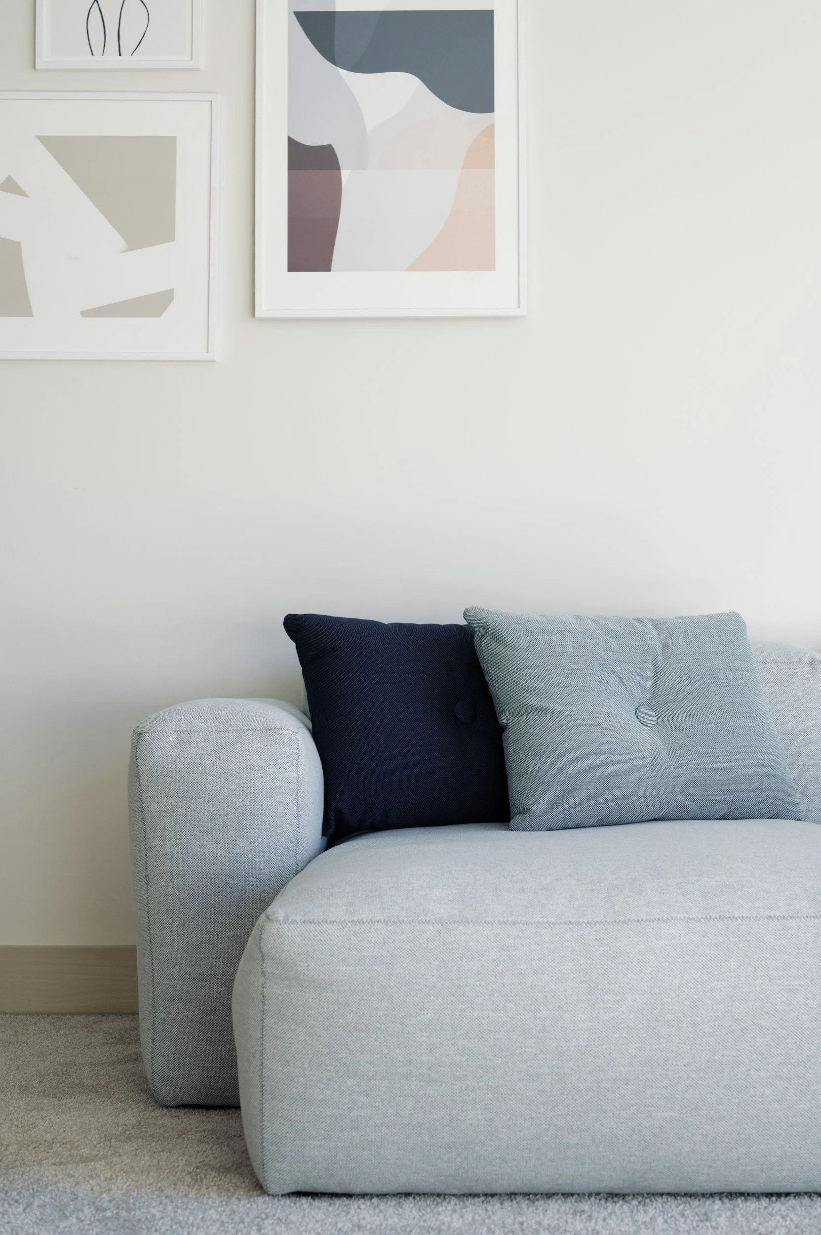 A light gray armchair with a rounded armrest and a cushioned seat, placed on a beige carpet in front of a white wall. Two pillows, one dark blue and one light gray, are on the chair. Two framed abstract art pieces hang on the wall behind the chair.