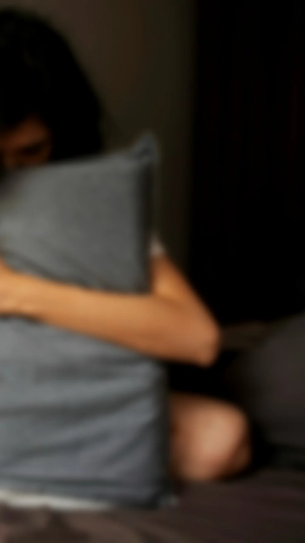 Blurred image of a person with dark hair sitting with a gray pillow or blanket.