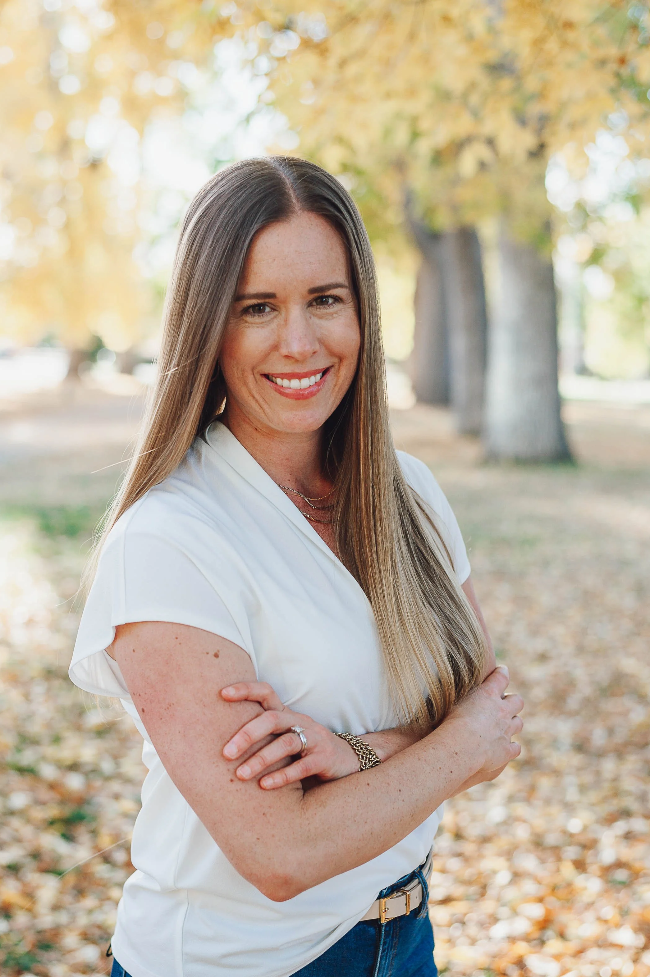 Kris Cristiano, LCSW, CSAT, wearing a white shirt and blue jeans, stands outdoors with her arms crossed, Denver autumn leaves on the ground, and trees in the background.
