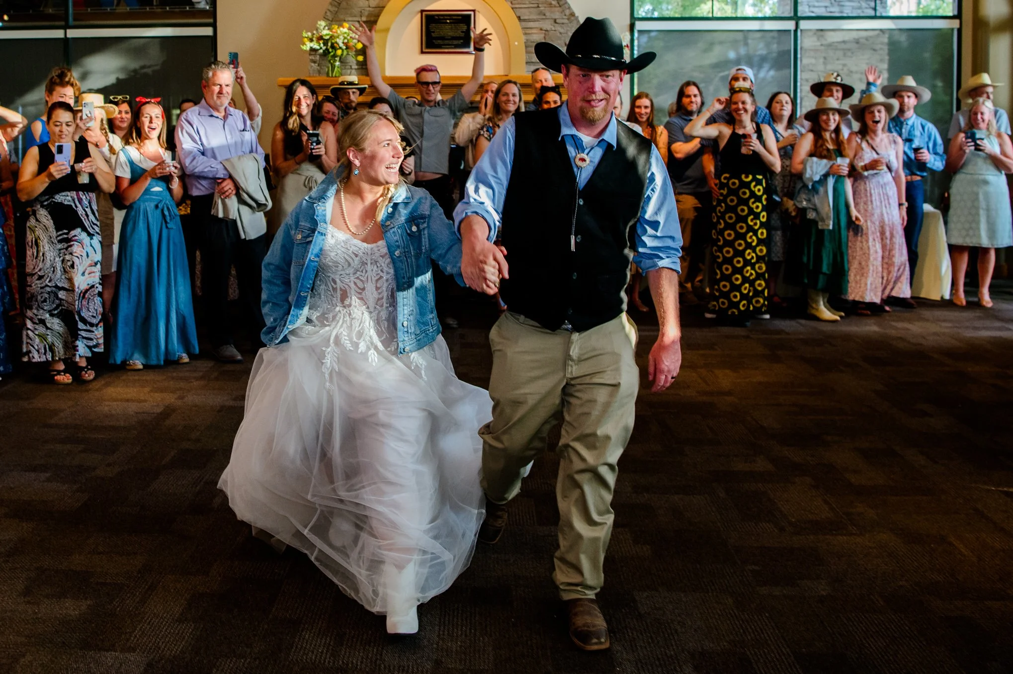 Bride and groom walk hand in hand into their reception as guests cheer and cameras flash.