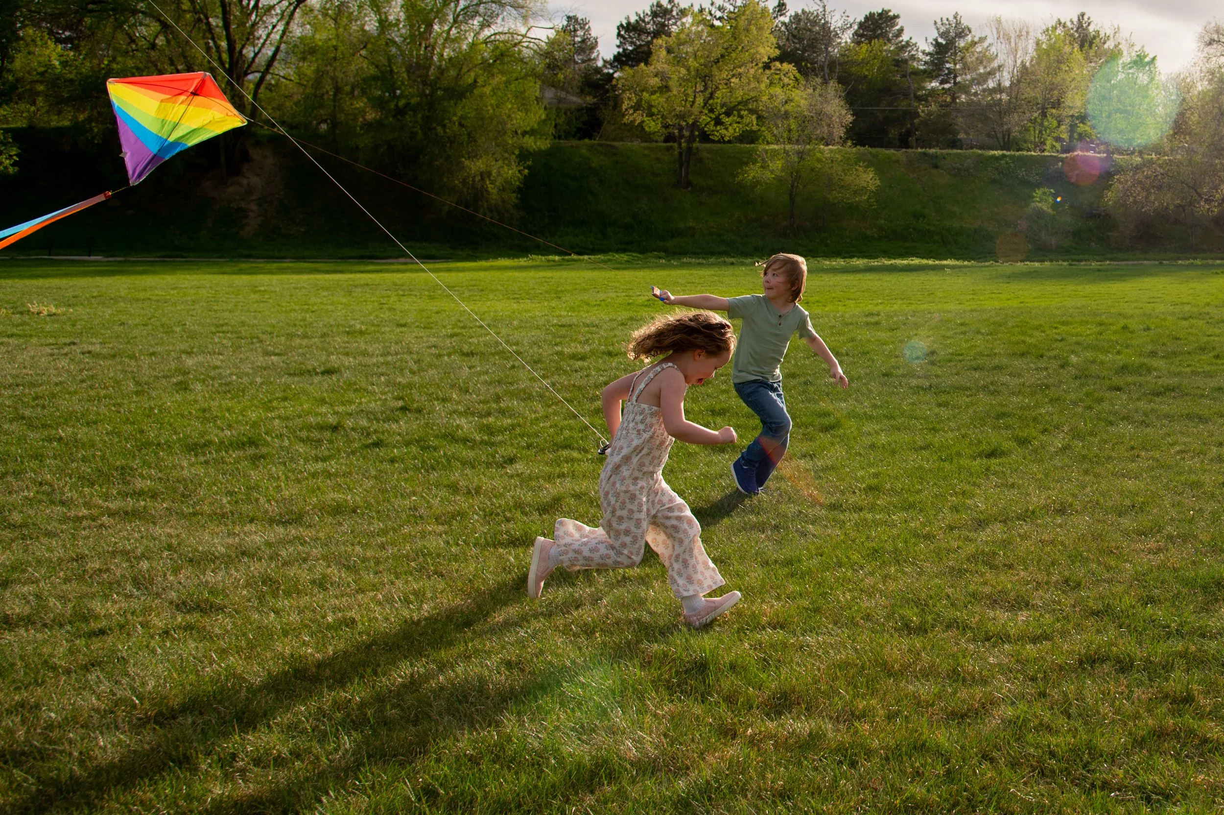 Two children, a boy and a girl, running on a grassy field chasing a colorful kite with a rainbow pattern as the sun sets behind trees in the background.