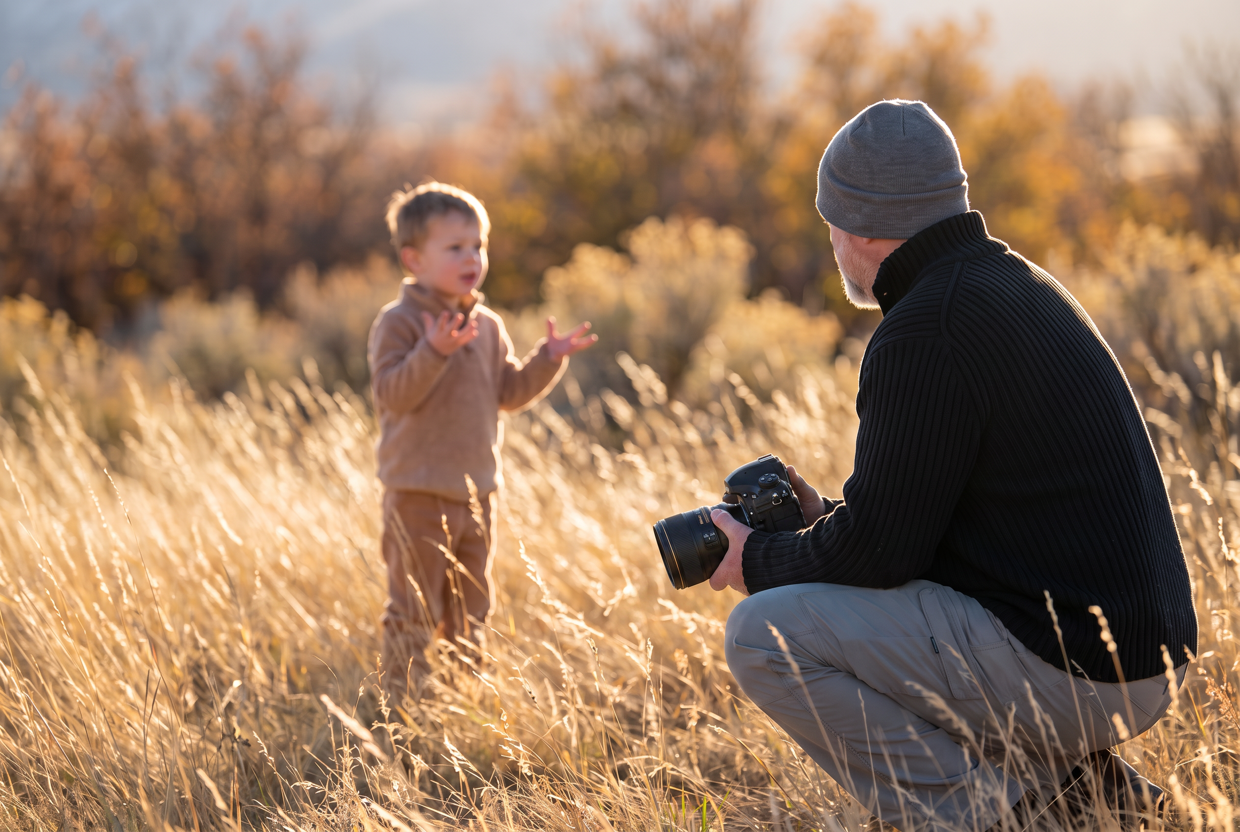 Brian Smyer crouches in tall golden grass with his camera lowered, listening to a young boy during a family session.