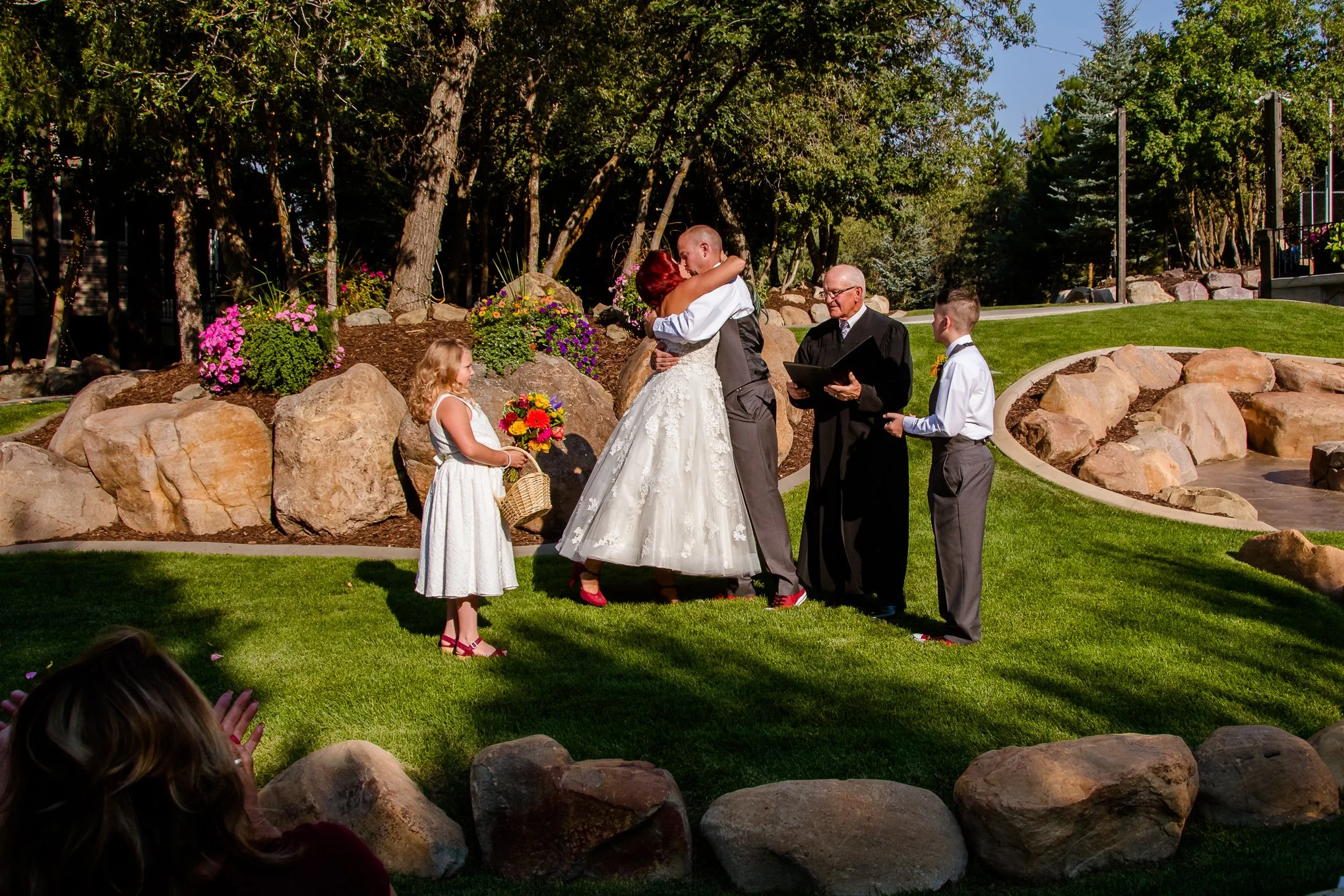 Bride and groom share their first kiss at an outdoor garden ceremony as an officiant and ring bearer look on.