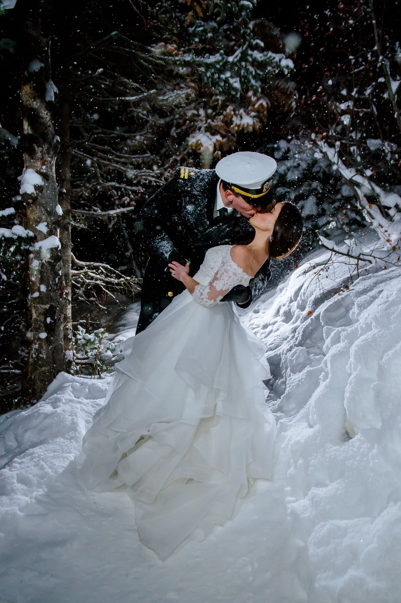 Bride and groom in military dress uniform share a kiss in falling snow surrounded by pine trees in Park City, Utah