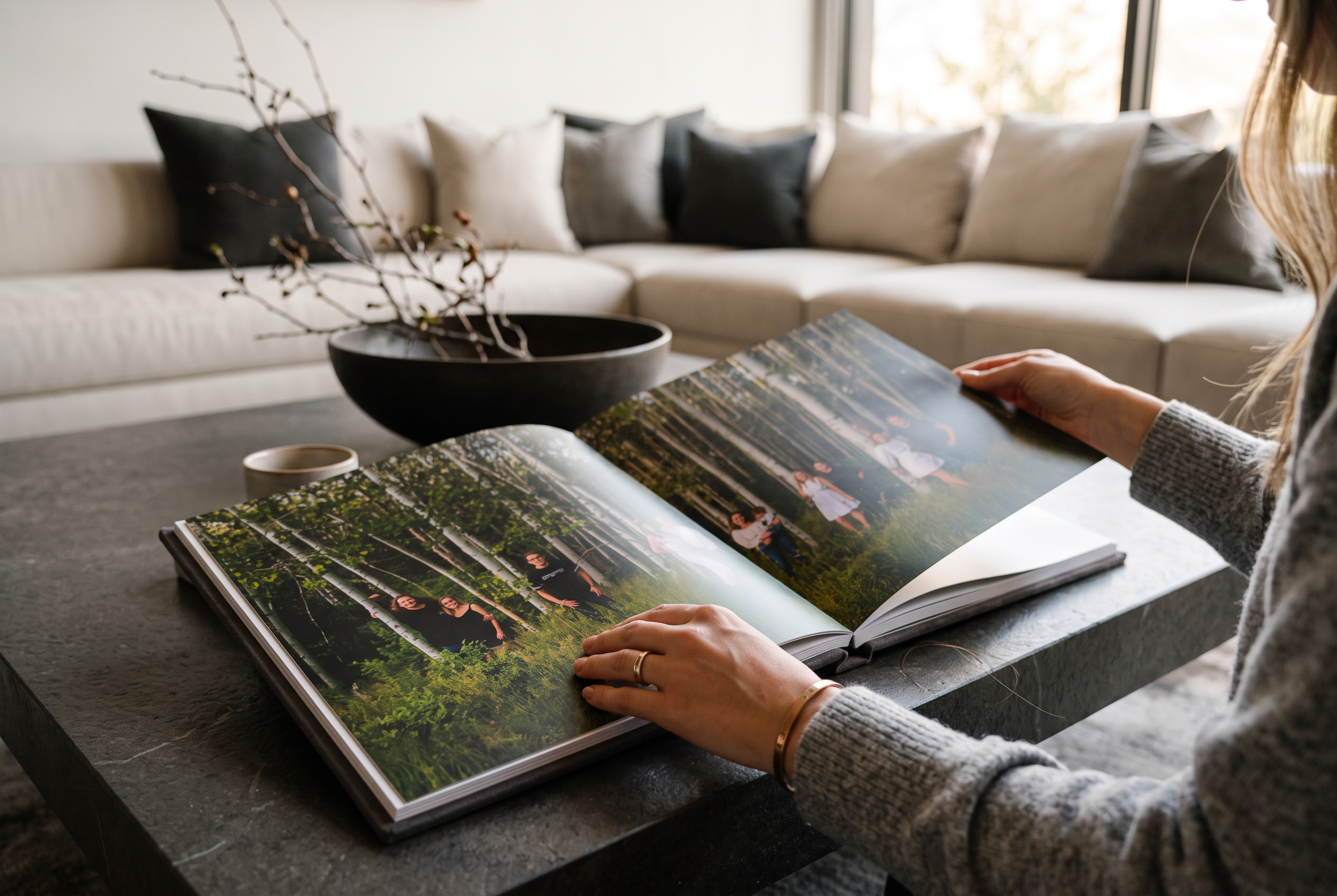 A person looking at a photo album on a table, with a black bowl and a small cup nearby. In the background, a light-colored sofa with black and white pillows and a window letting in natural light.