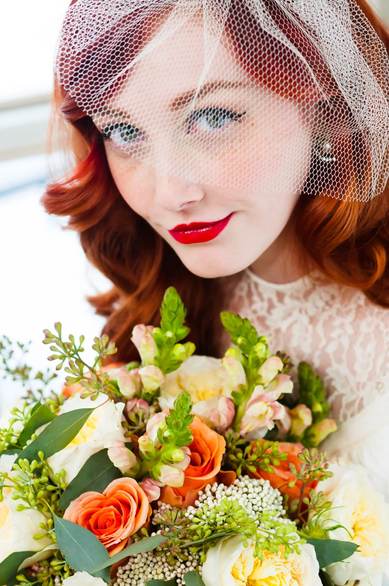 Close bridal portrait featuring a birdcage veil, red lipstick, and a citrus-toned bouquet.