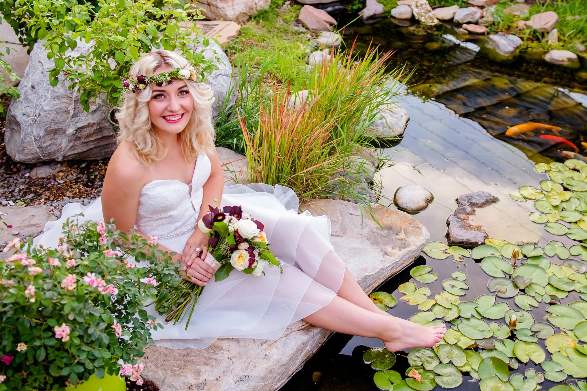 Bride in a flower crown sits barefoot beside a koi pond holding a wildflower bouquet.