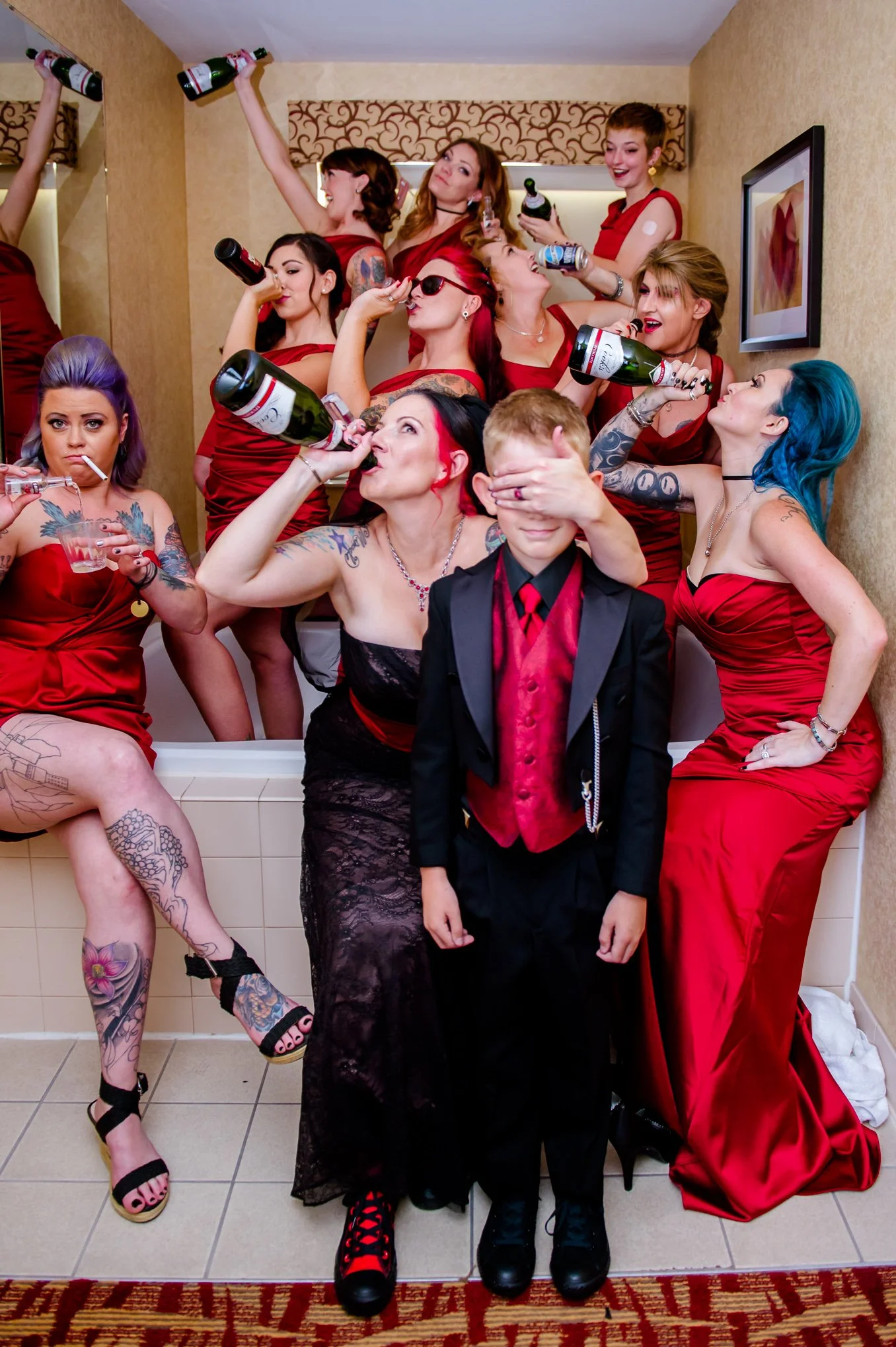A bridal party in red dresses celebrates with drinks in a hotel bathroom while a young boy in a black and red tuxedo covers his eyes.