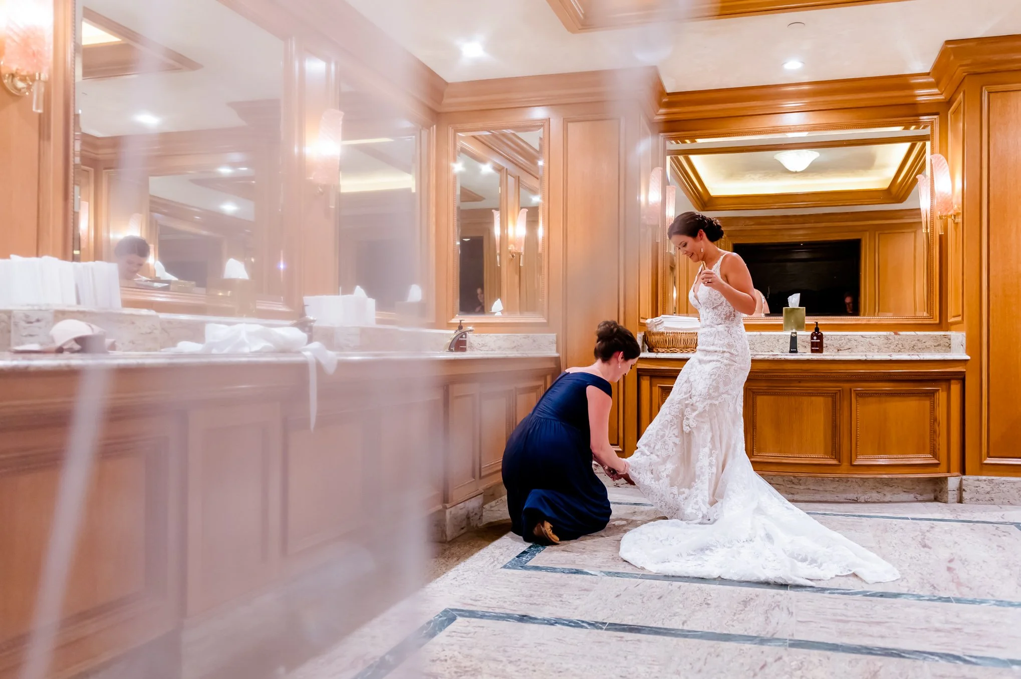 A bridesmaid adjusts the bride's train in a hotel powder room while getting ready before the ceremony.