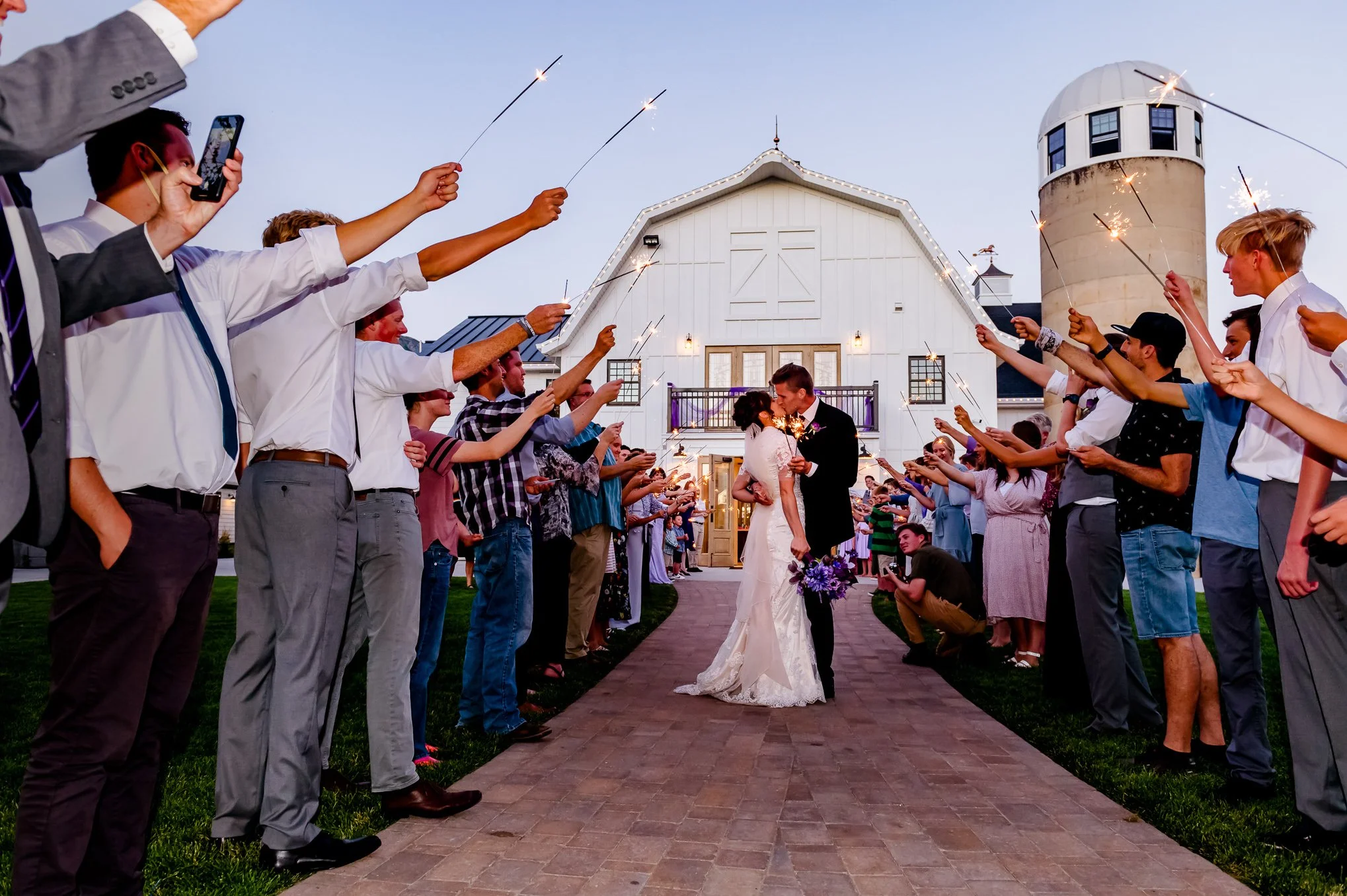 Bride and groom kiss during their sparkler exit in front of a white barn at Walker Farms at dusk.