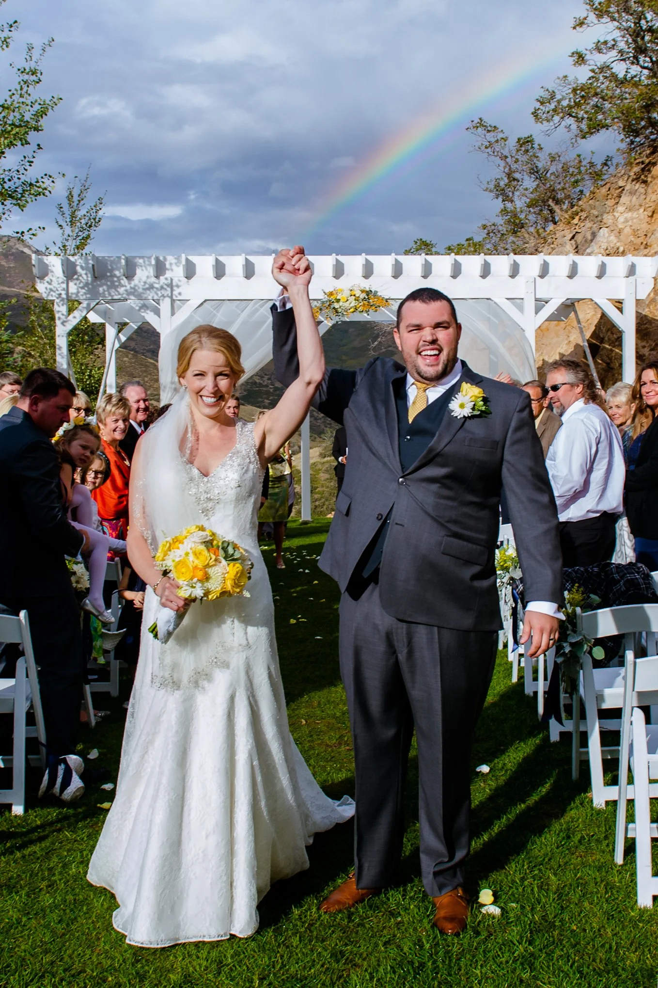 Newlyweds raise their hands in celebration under a rainbow as they walk back down the aisle at Louland Falls