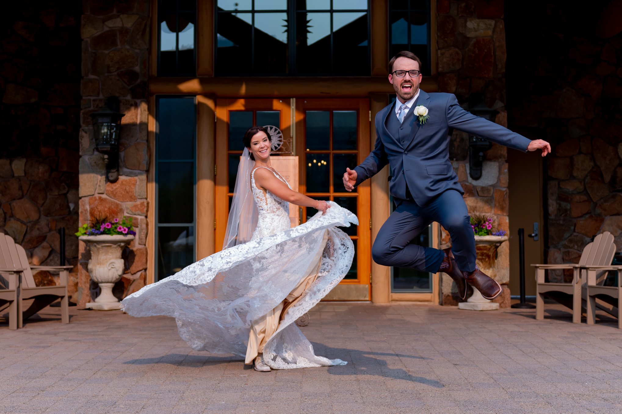 Groom leaps in the air while his bride laughs and twirls her dress outside their venue.