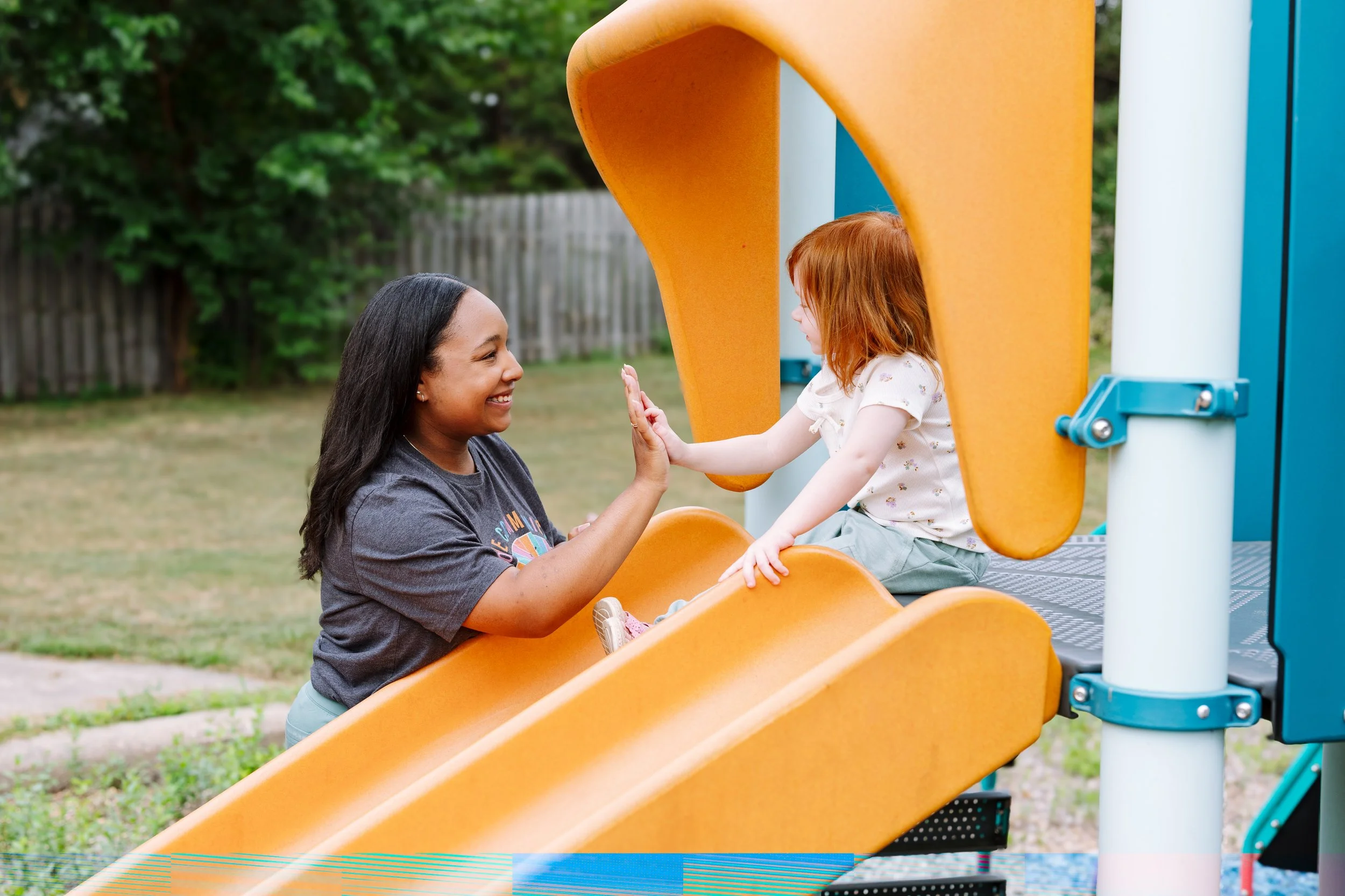 A woman and a young girl smiling and giving each other a high five while sitting on a yellow playground slide.