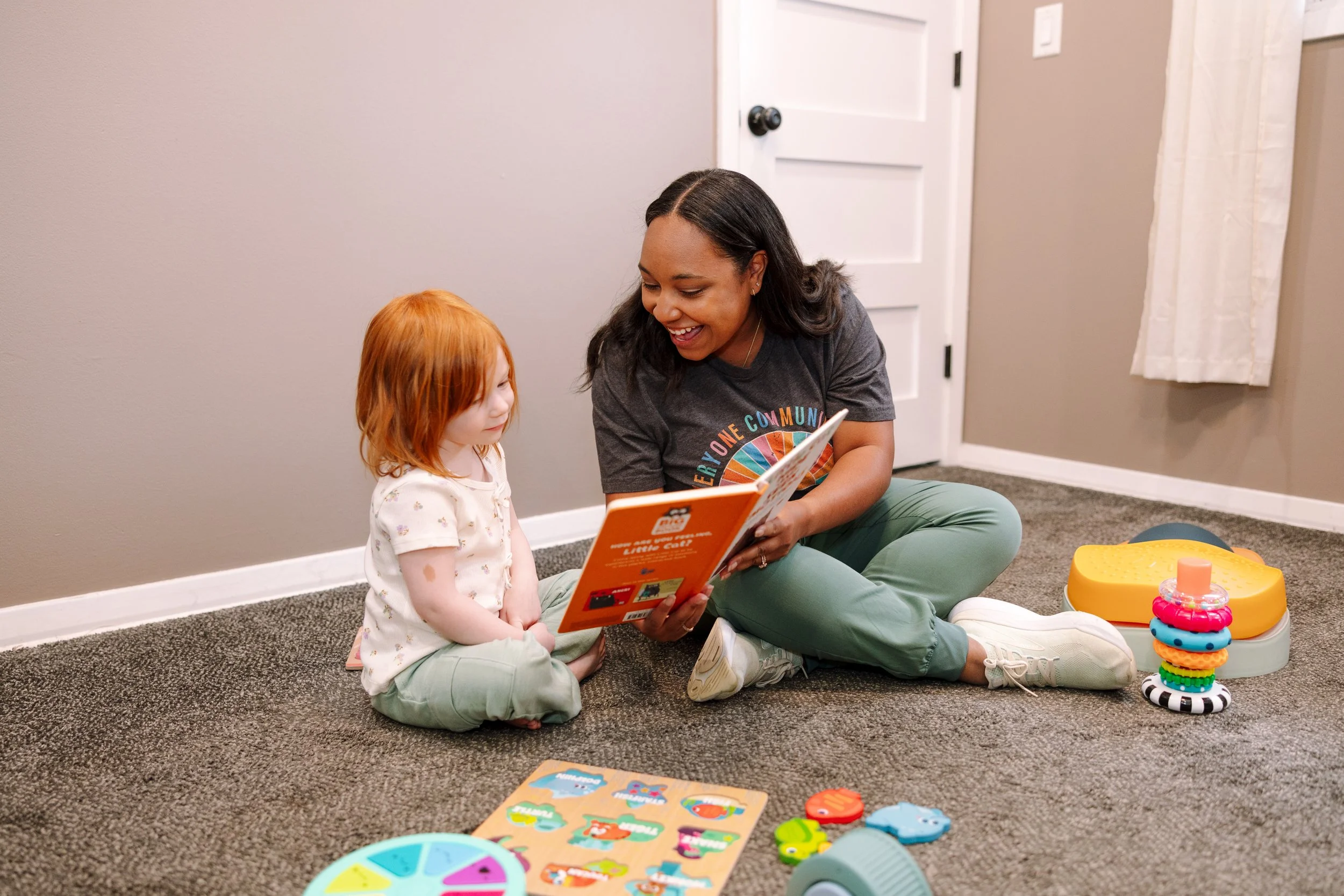 A young woman with dark hair smiling and reading a book to a little girl with red hair sitting on the carpet in a cozy room with toys on the floor.
