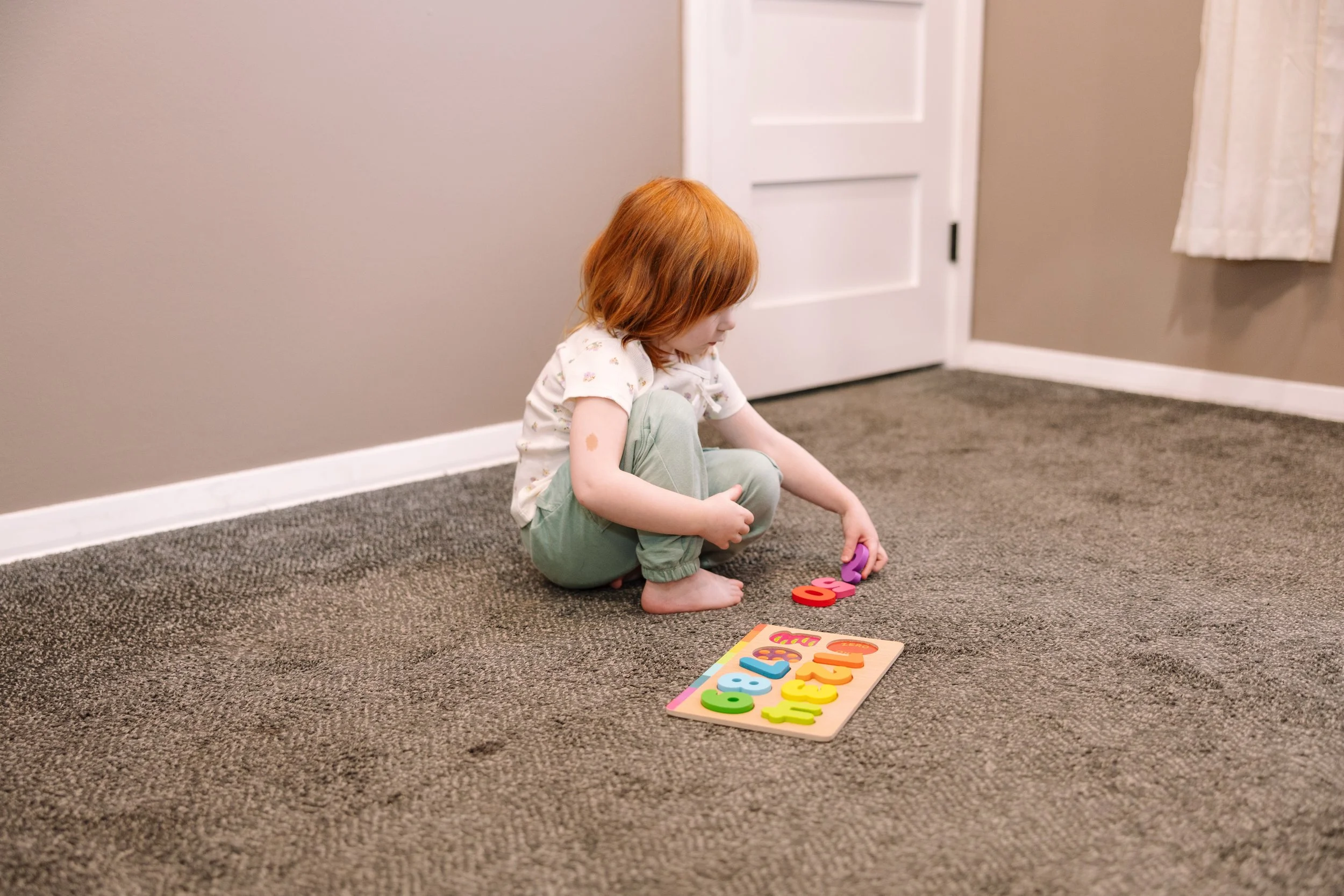 A young girl with red hair sitting on a carpeted floor, playing with colorful foam numbers and a wooden puzzle board with numbers on it in a room with beige walls, white door, and a curtain.